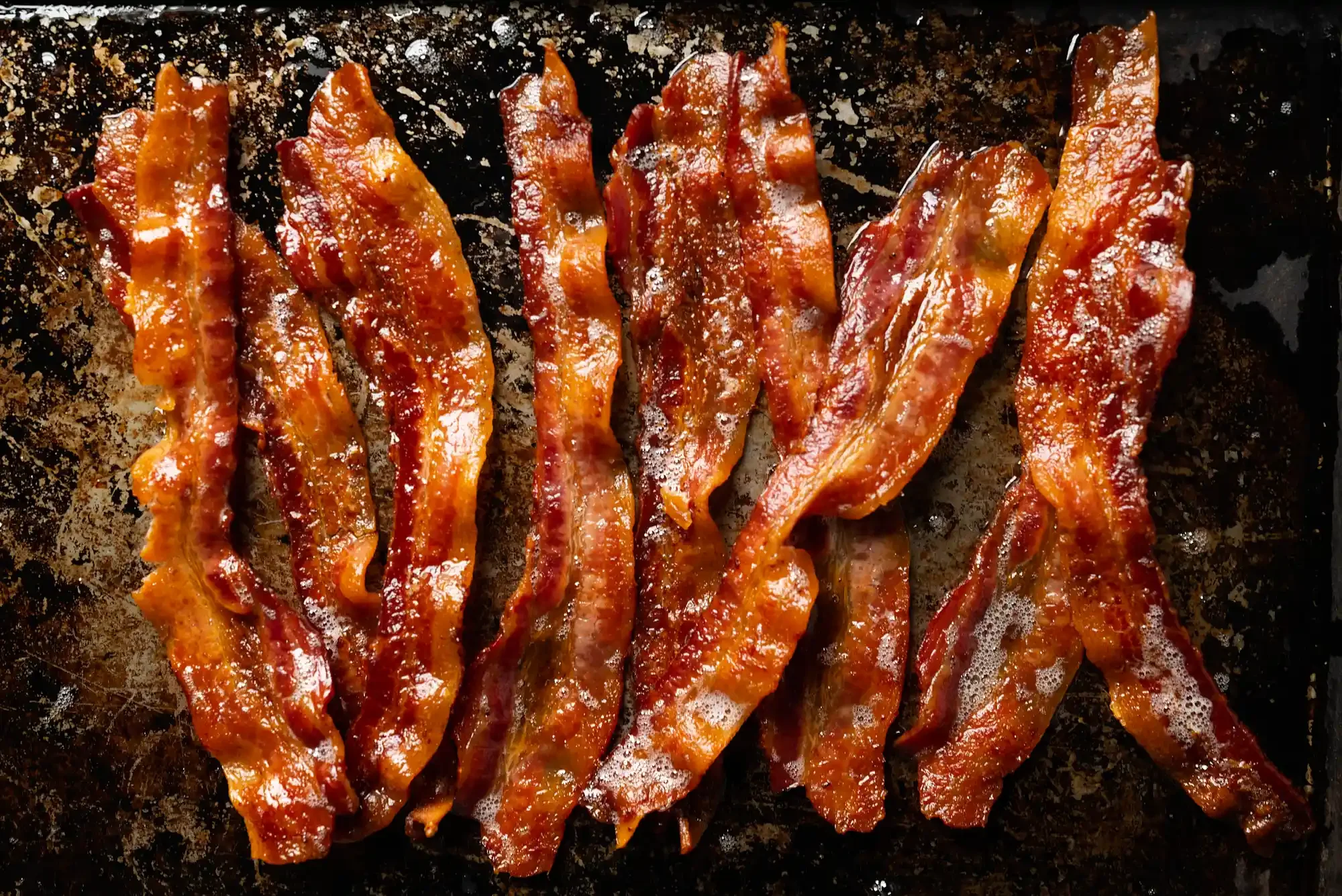 Bacon strips on a cooking surface, sizzling and slightly glazed. By Philadelphia, PA food photographer & food stylist team Bartholomew Studio.