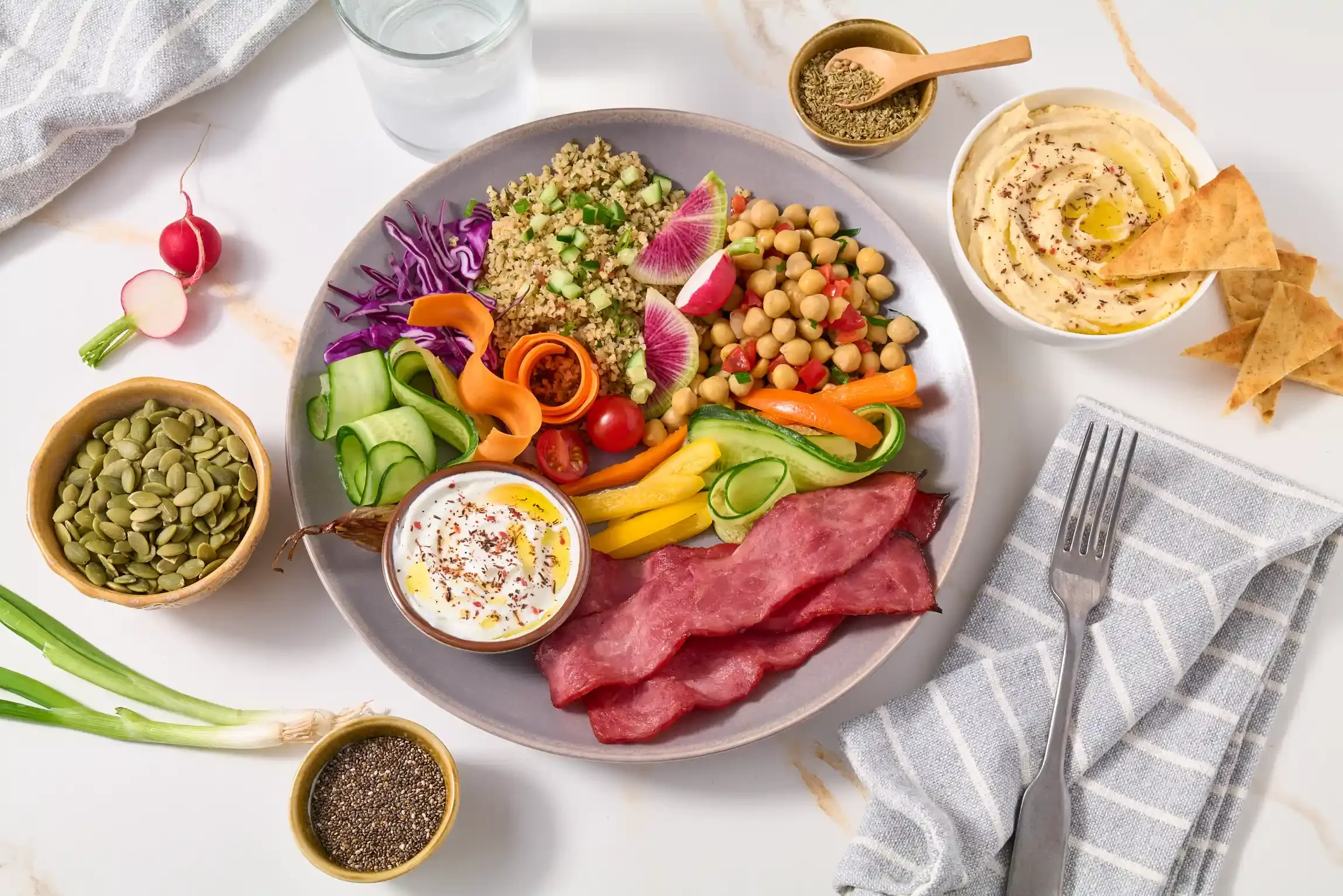 A plate of colorful vegan food with chickpeas, quinoa, fresh vegetables, and sliced fruit, accompanied by bowls of hummus, guacamole, pumpkin seeds, chia seeds, and a side of pita chips. By Philadelphia, PA food photographer & food stylist team.