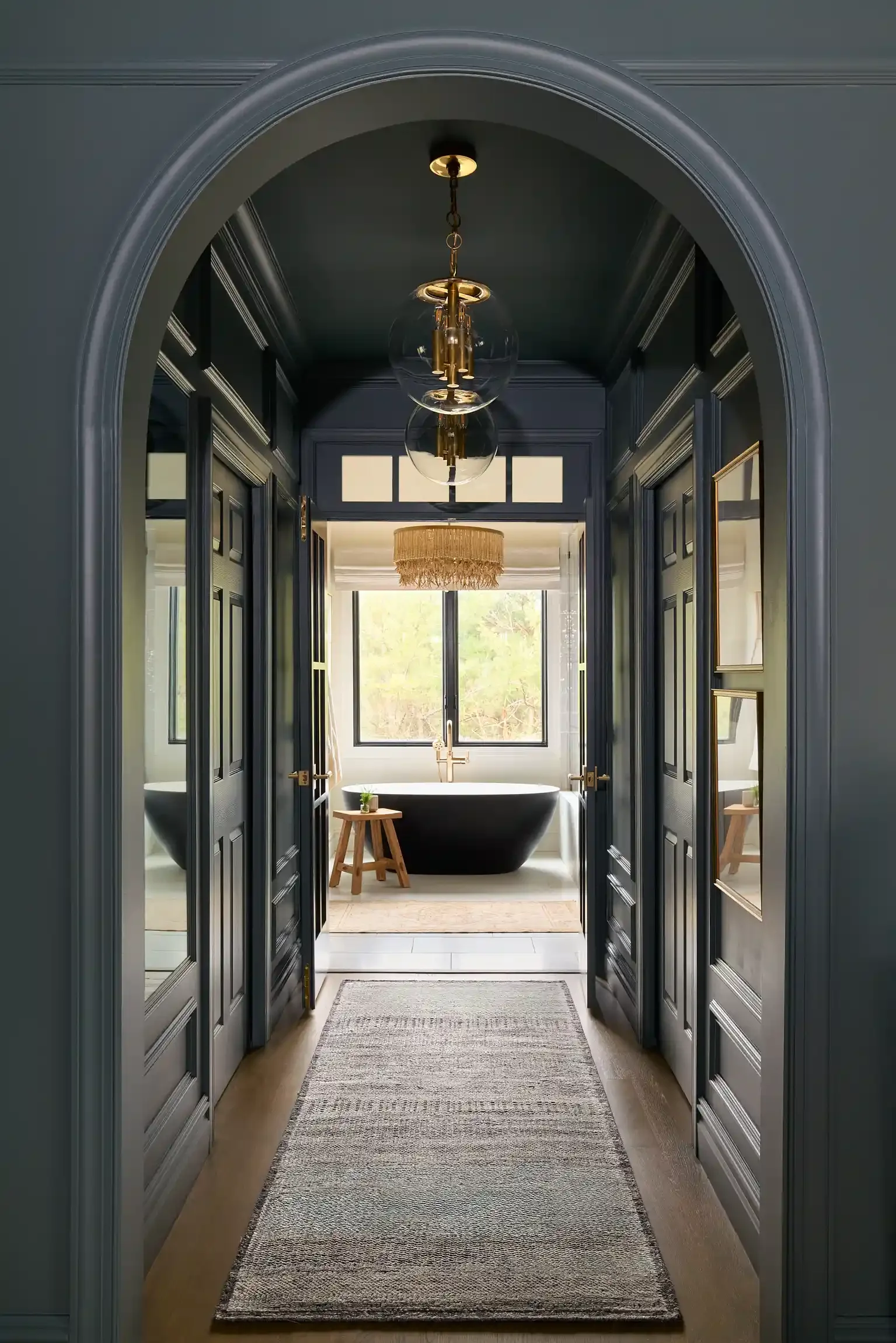 Hallway leading to a bathroom with a freestanding bathtub, large window, a small wooden stool, and a ceiling light fixture. By New Jersey interior photographer.