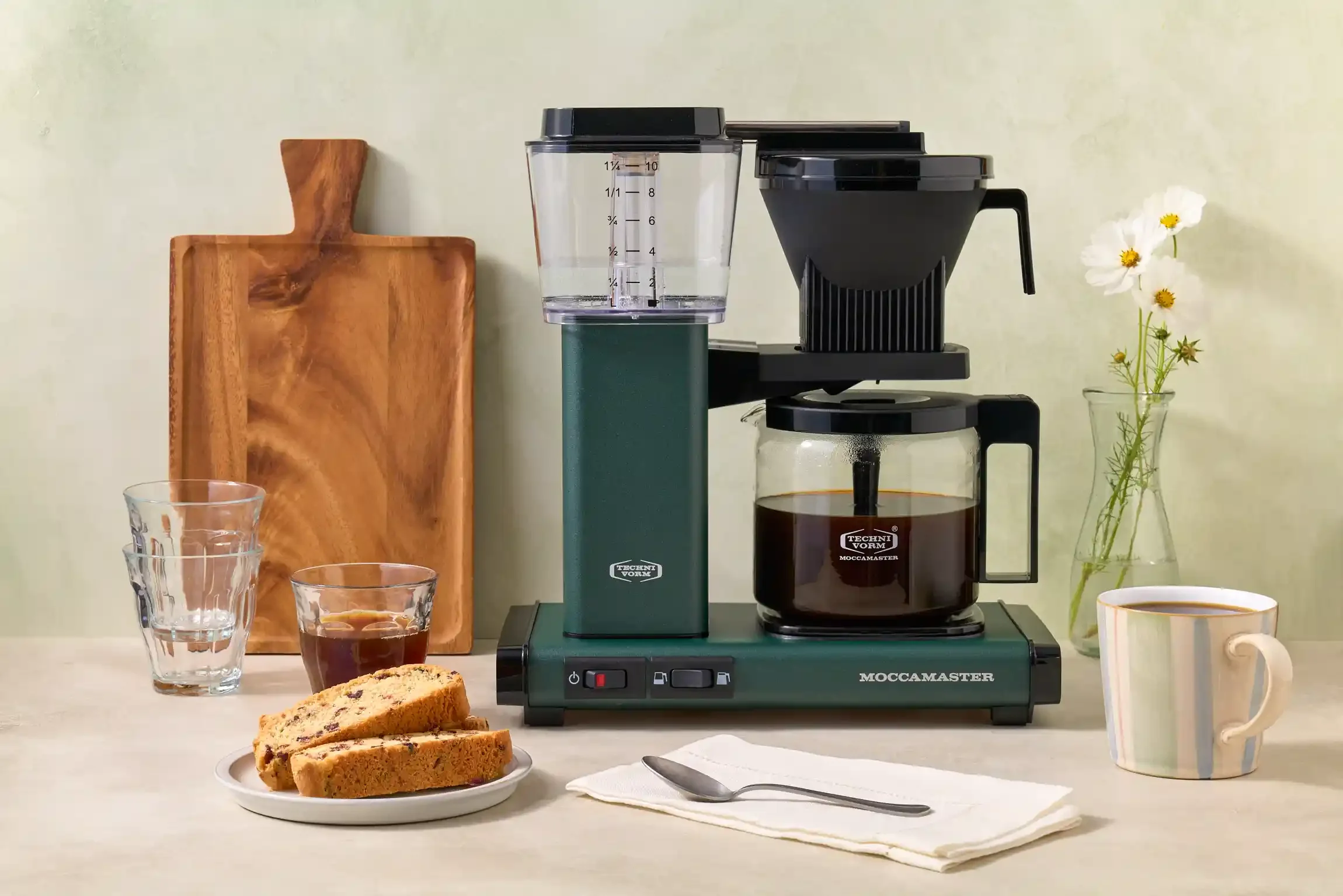A Moccamaster coffee maker brewing coffee on a kitchen countertop with a plate of biscotti, a cup of coffee, a spoon on a napkin, a small flower vase with white flowers, and a wooden cutting board. By Philadelphia, PA & NJ coffee drink photographer. 