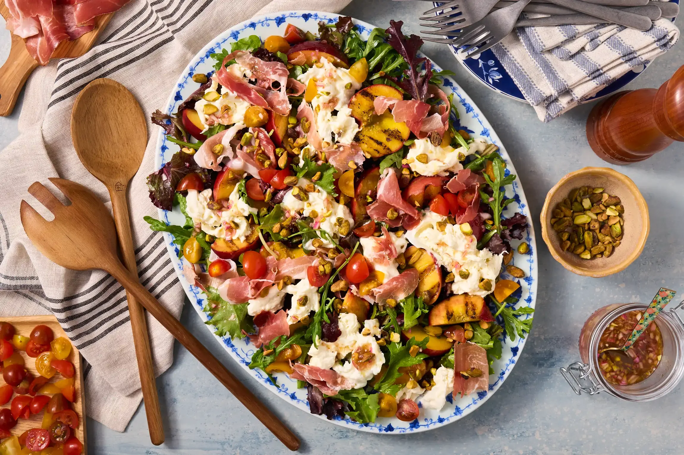 A colorful mixed salad on a blue and white patterned plate, with cherry tomatoes, greens, cheese, prosciutto, grilled peaches, and pistachios. By Philadelphia, PA & NJ Food Photographer & Stylist Team.