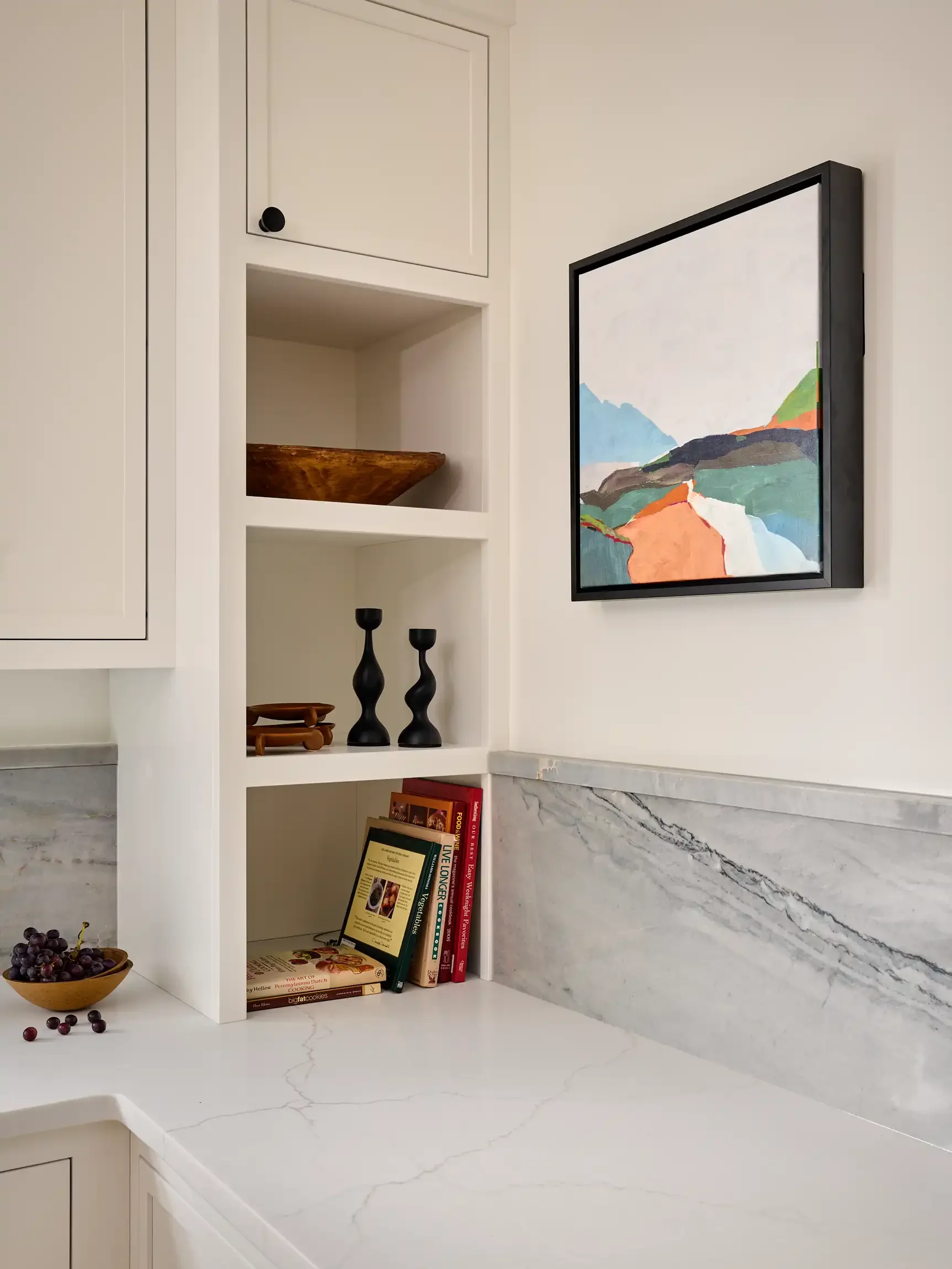 A kitchen countertop with white cabinetry with black knobs and a framed landscape painting on the wall. By Restart Homes.