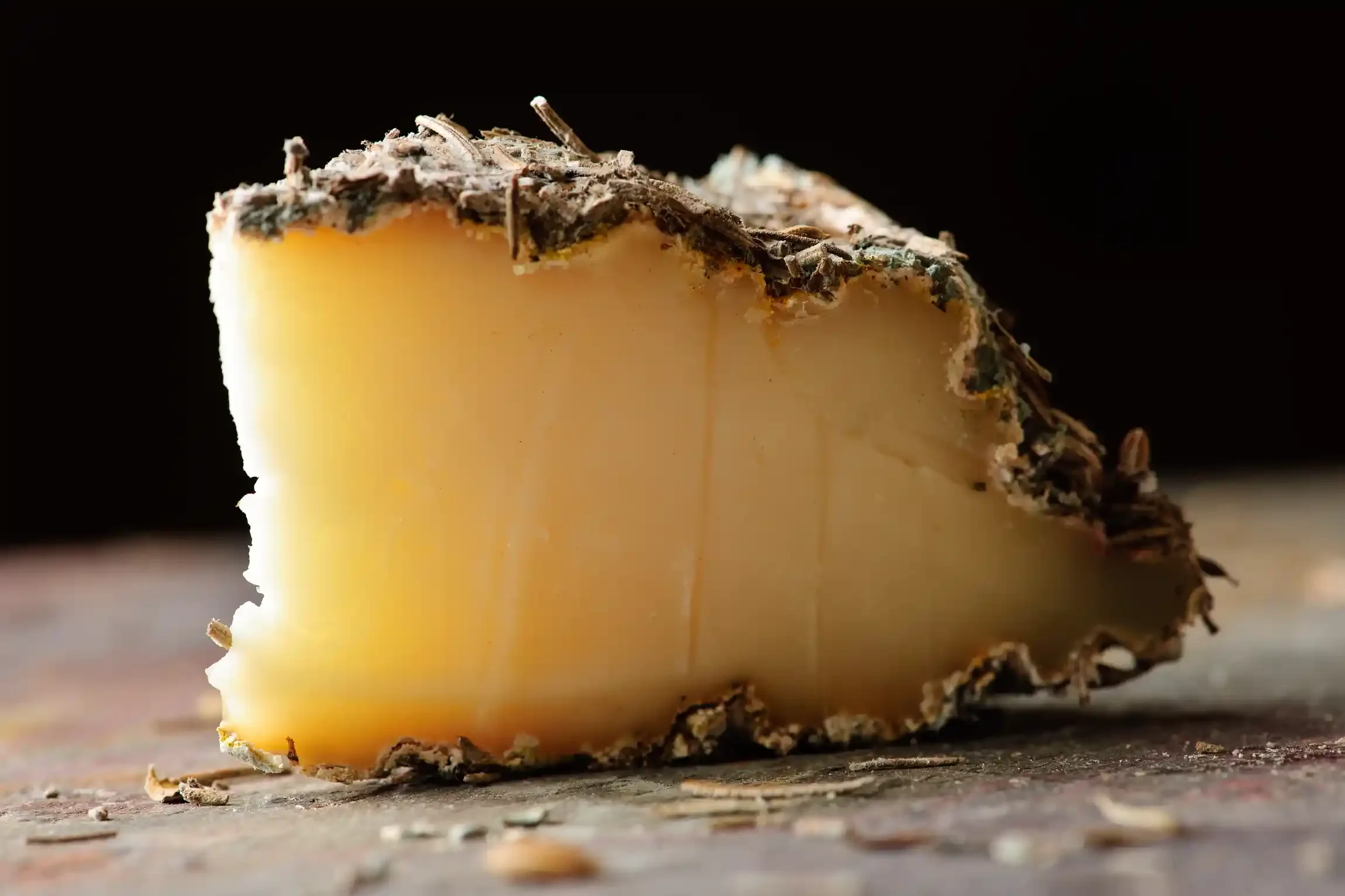 Close-up of a wedge of French Brin d'Amour cheese with a dark rind, sitting on a wooden surface, against a black background. By Philadelphia, PA & NJ food photographer & food stylist team Bartholomew Studio.