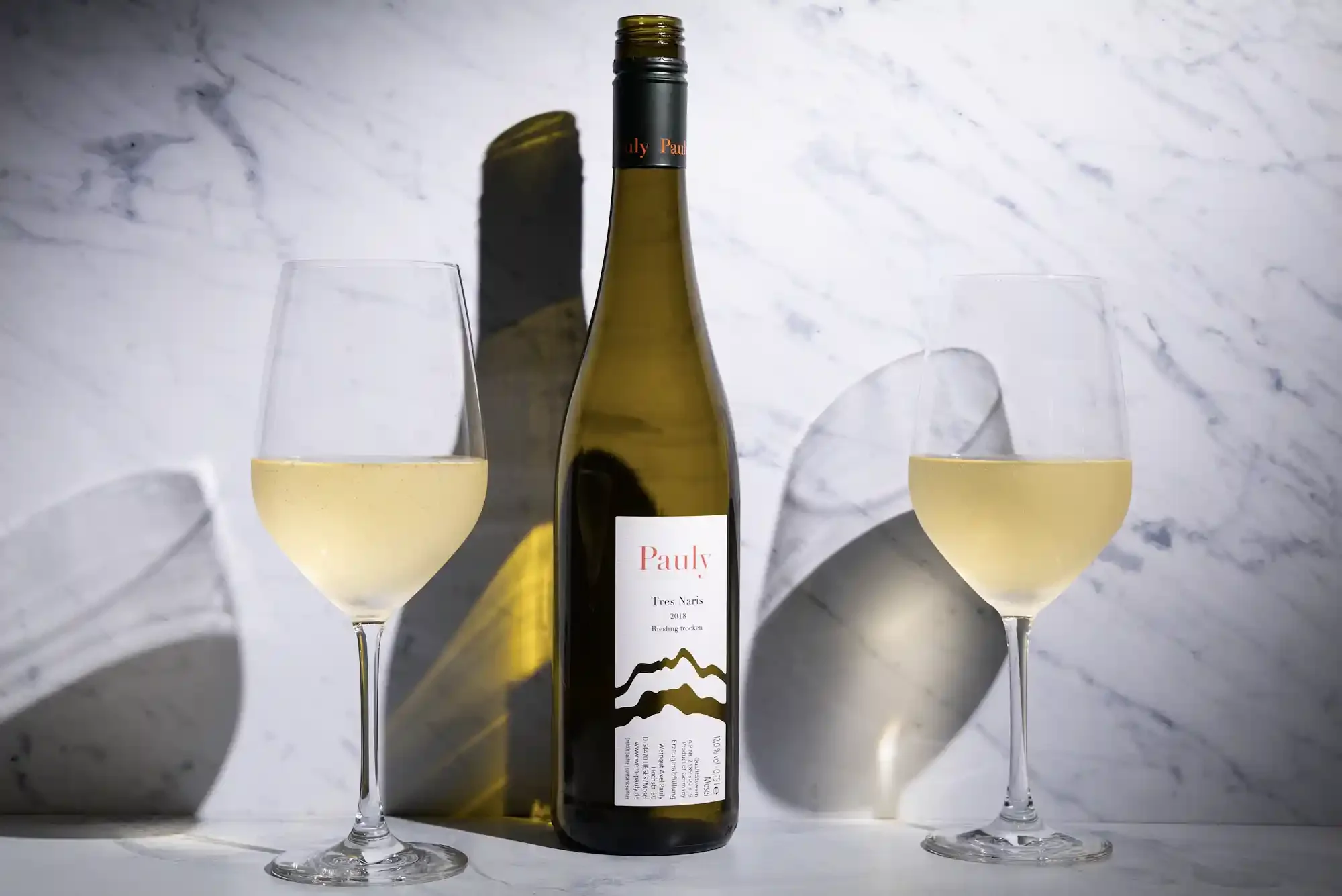 A bottle of white wine placed between two filled wine glasses on a surface, with a marble wall in the background and shadows cast on the wall. By Philadelphia, PA & New Jersey drink photographer & drink stylist team Bartholomew Studio.