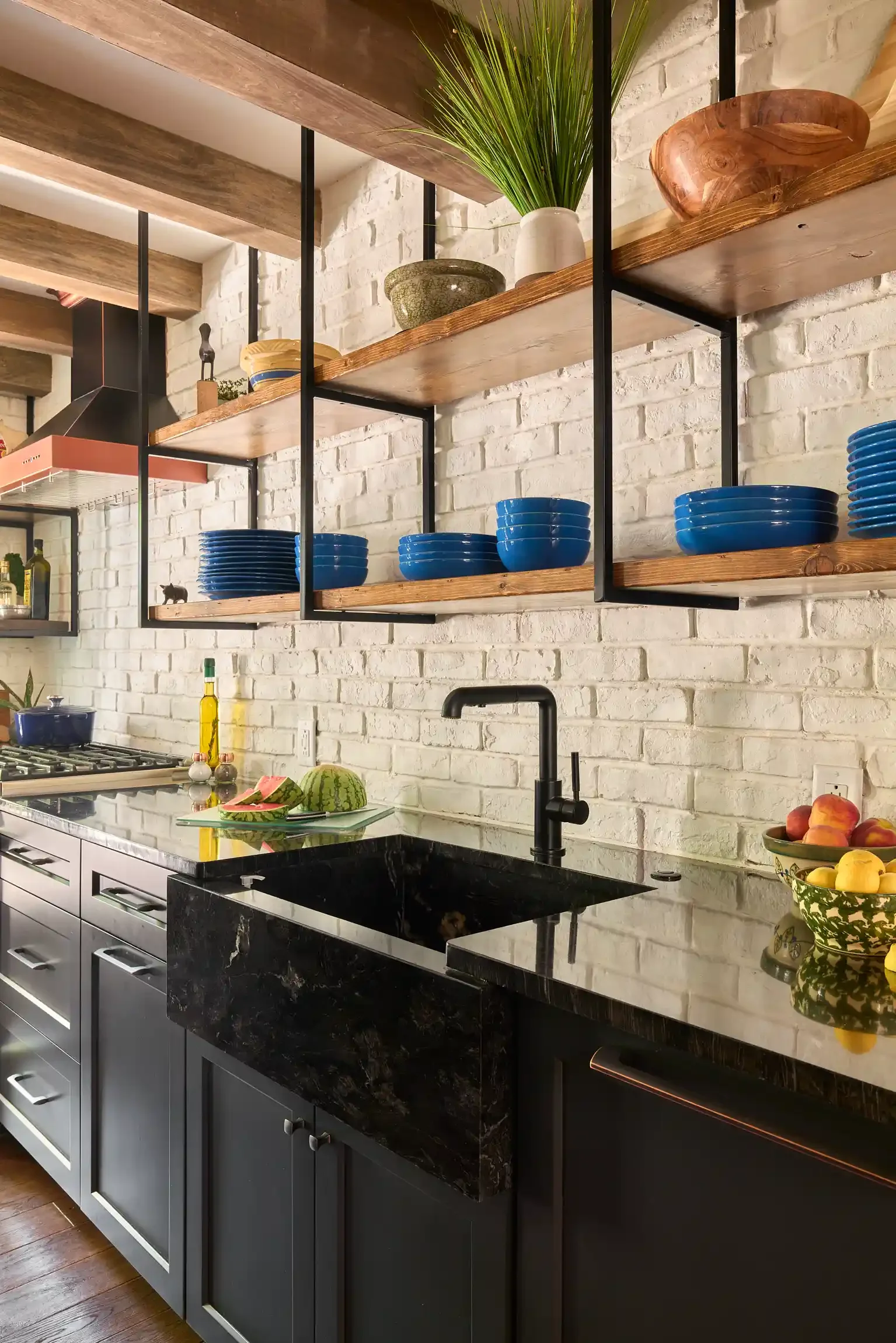 Kitchen with white brick wall, black countertop, open wooden shelves holding blue bowls, green and beige decorative bowls, and green potted plant. Black sink with peaches and watermelons on the counter. By Bucks County, PA interior photographer.