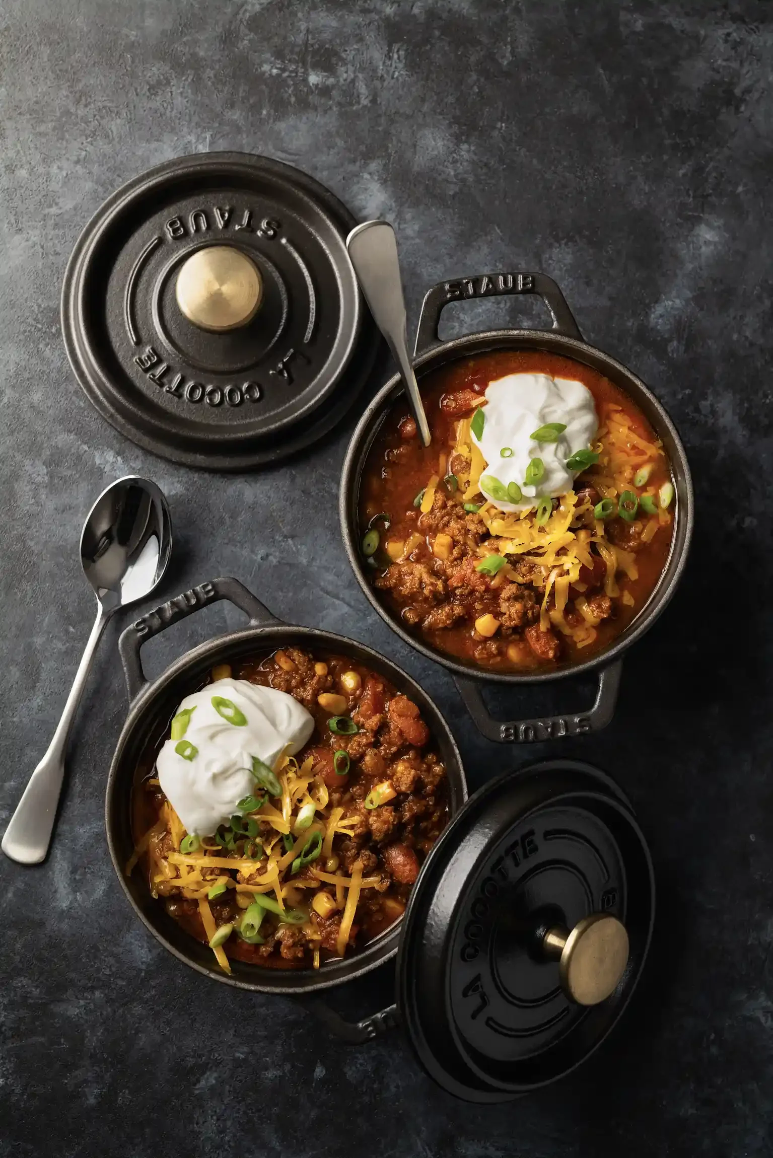 Two small cast iron skillets filled with chili topped with shredded cheese, sour cream, and chopped green onions, placed on a dark surface. By New York food photographer & food stylist team Bartholomew Studio.