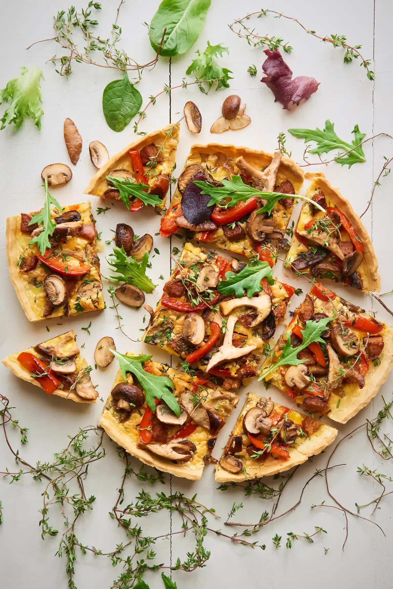 Vegetarian mushroom and red pepper pizza with herbs on top, scattered green and purple leaves, and small sprigs of thyme on a white wooden surface. By Philadelphia, PA & NJ food photographer & food stylist team Bartholomew Studio.