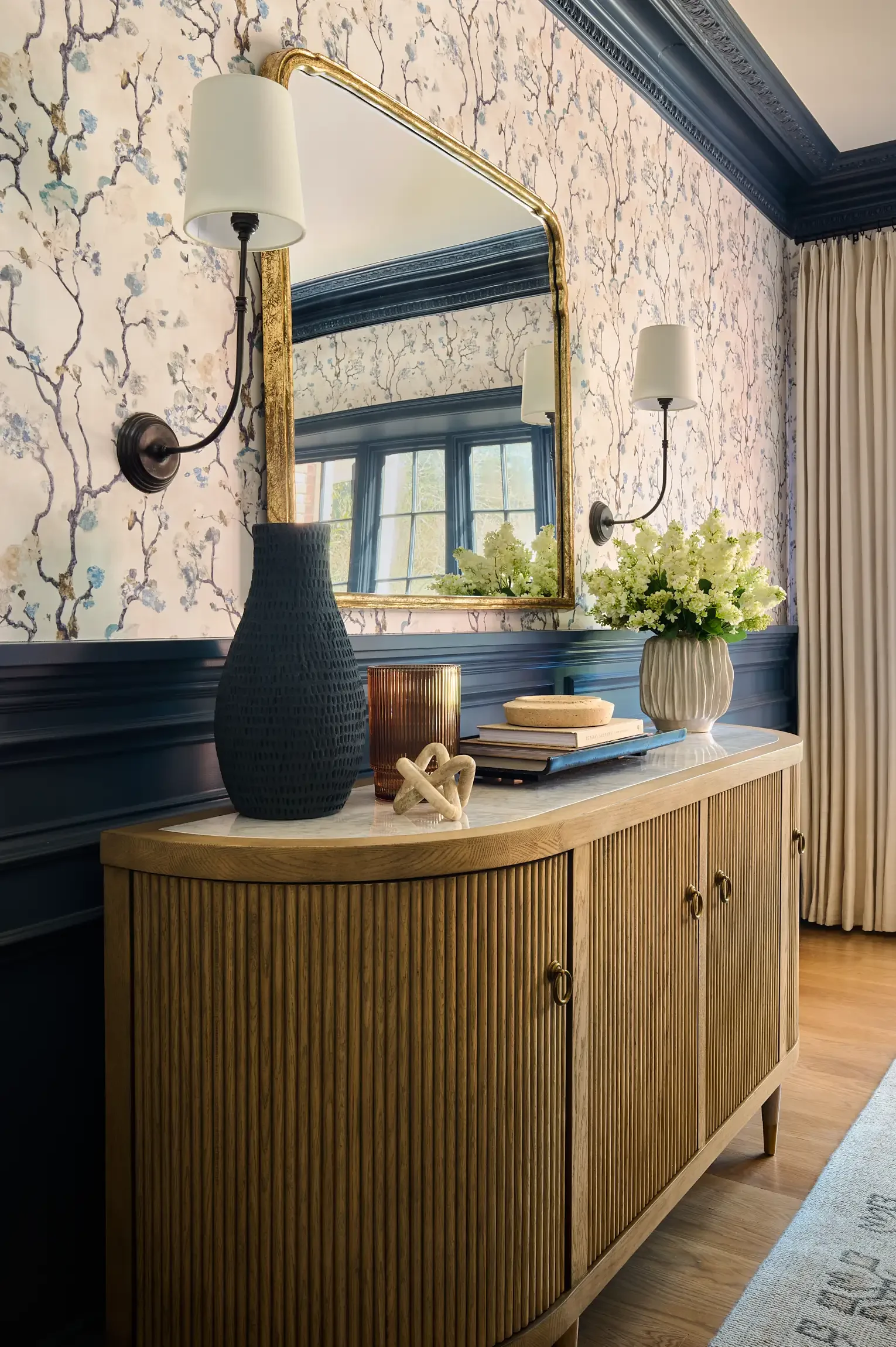 A wooden sideboard with a marble top, decorated with a large black textured vase, a smaller amber-colored vase, a stack of books topped with decorative objects, and a large white vase with flowers. By interior design photographer Paul Bartholomew.