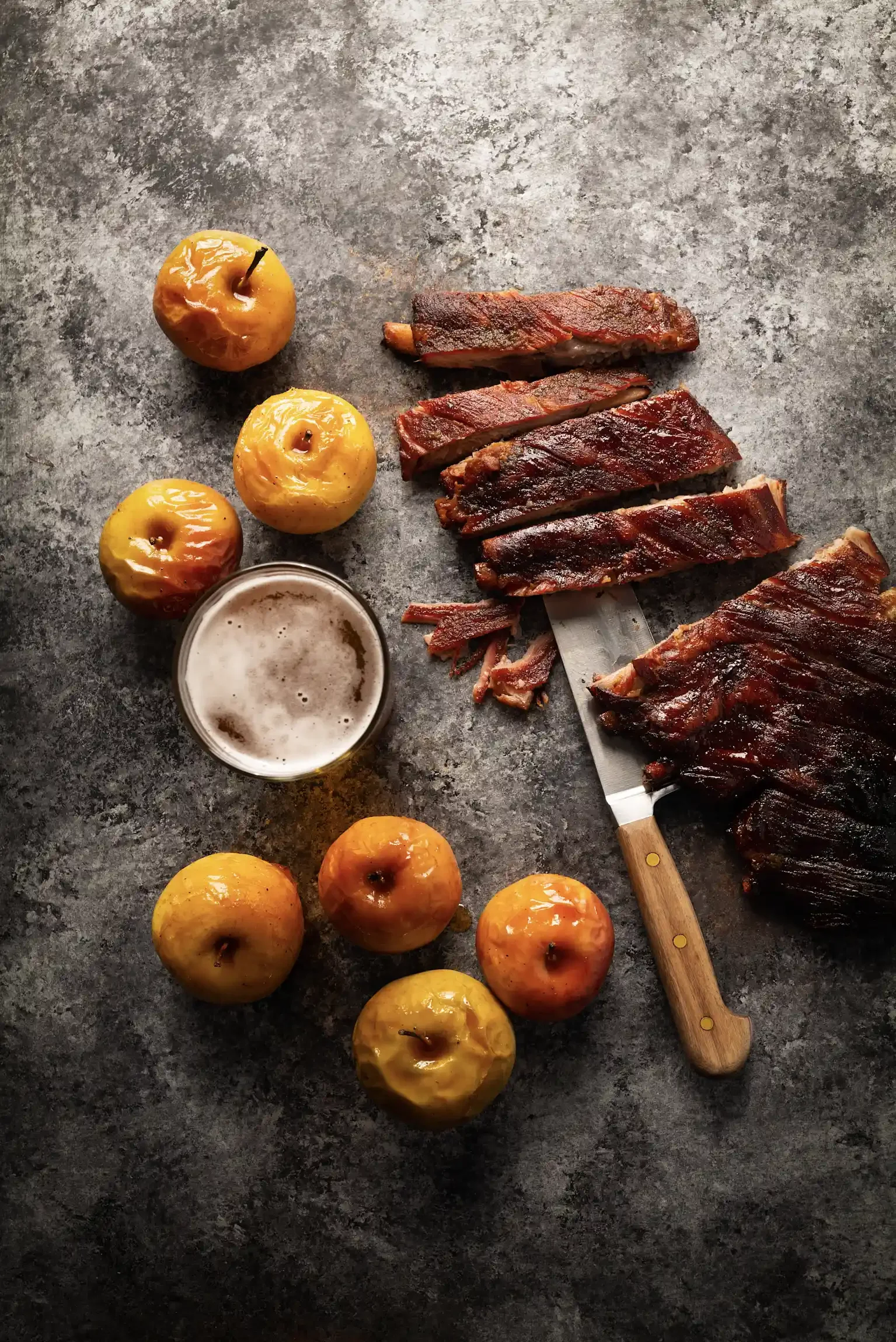 Ribs, apples, and a glass of beer on a gray textured surface. By New Jersey food photographer & food stylist team Bartholomew Studio.