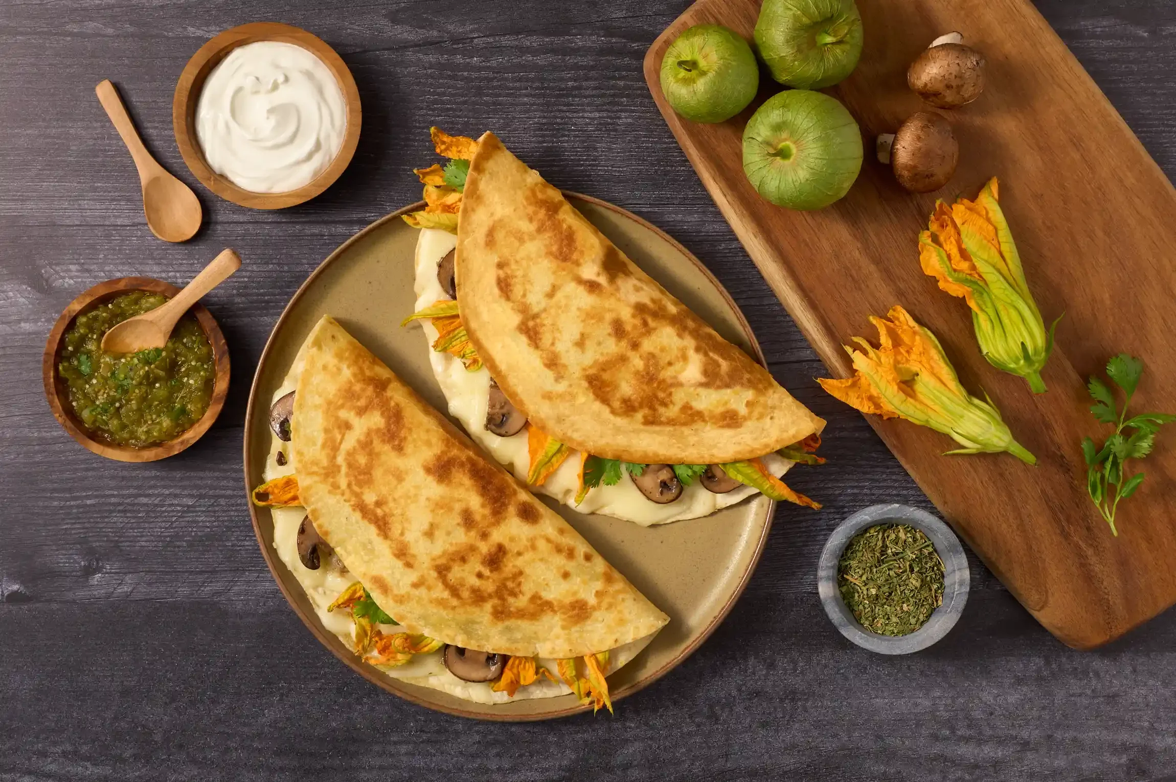 Two quesadillas filled with mushrooms, squash blossoms, and cheese on a beige plate, surrounded by small bowls of sour cream, salsa verde, and herbs. By Philadelphia, PA & NJ food photographer & food stylist team Bartholomew Studio.