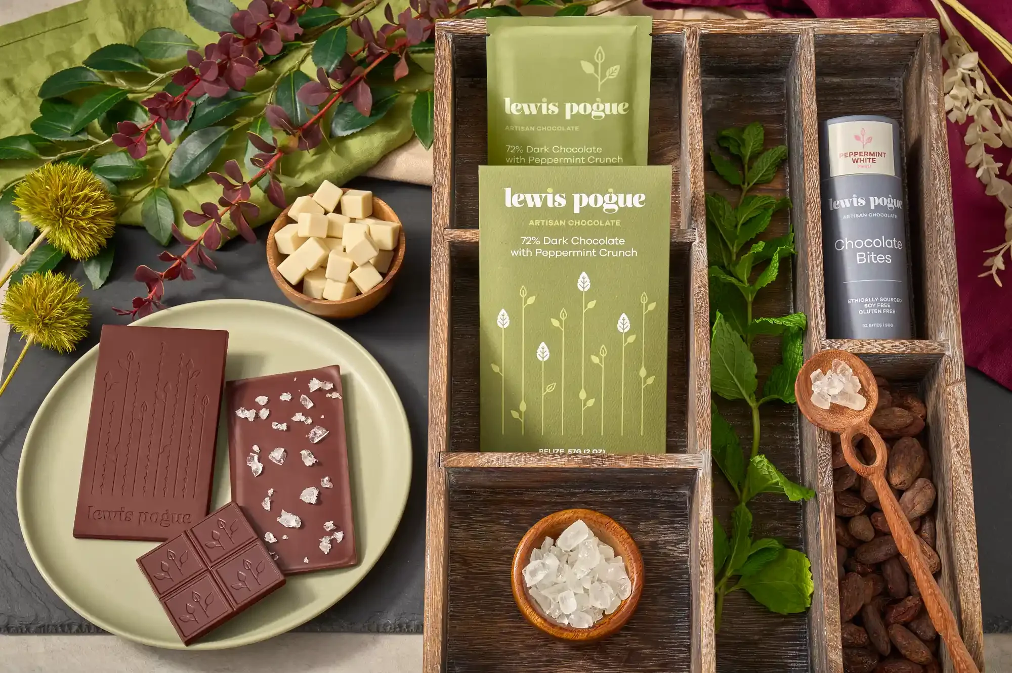 Assorted chocolates and ingredients on a table, including dark chocolate with peppermint crunch, white chocolate, sea salt, and cocoa beans and decorative flowers. By Philadelphia, PA chocolate photographer & food stylist team Bartholomew Studio.