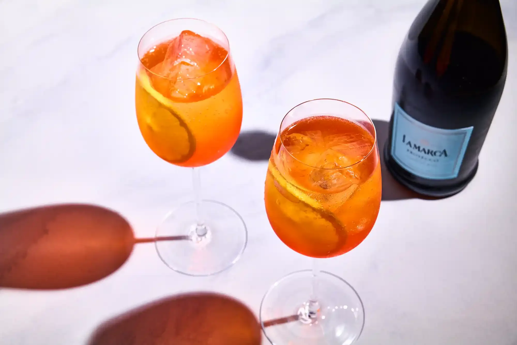 Two glasses of Aperol spritz with ice, slices of orange and placed on a white surface. By Pennsylvania drink photographer & drink stylist team Bartholomew Studio.