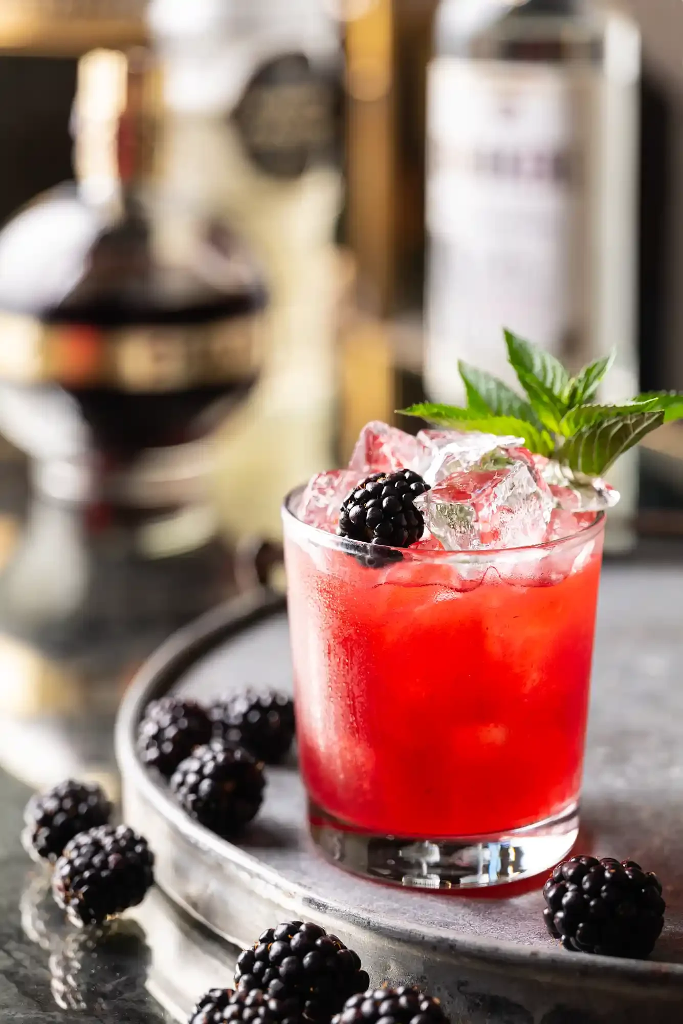 A glass of blackberry bramble cocktail with ice, garnished with blackberries and mint leaves, on a gray surface with additional blackberries around it. By Philadelphia, PA & NJ drink photographer & drink stylist team Bartholomew Studio.