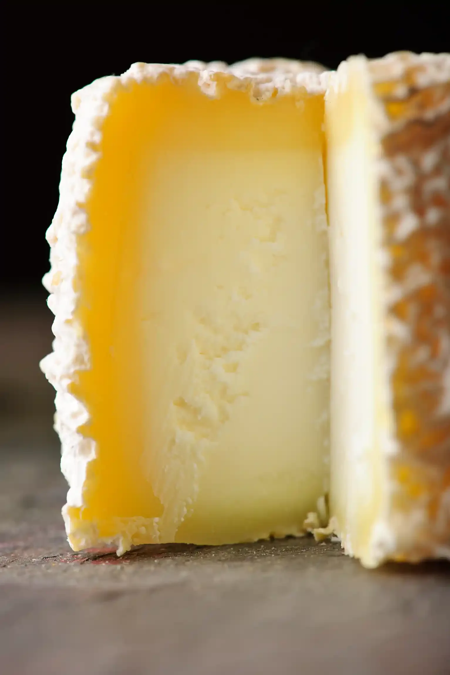 Close-up of a wedge of goat cheese with a textured rind, showing a creamy, yellow interior. By Philadelphia, PA & NJ cheese photographer & food stylist team Bartholomew Studio.