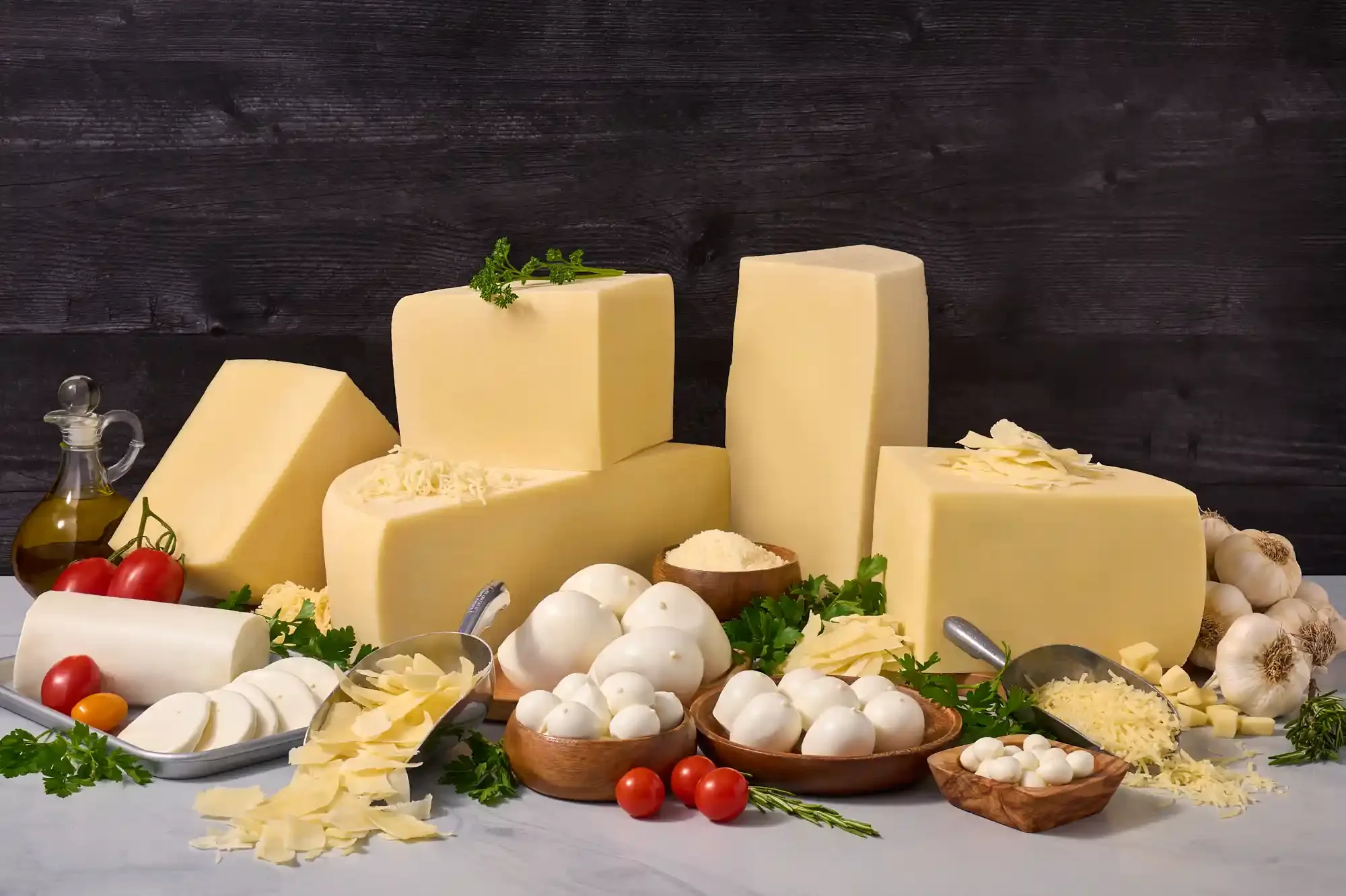 Various types of cheese including blocks and slices, surrounded by tomatoes, garlic, herbs, and other dairy products on a kitchen table. By PA, NJ & NY cheese photographer & stylist team Bartholomew Studio.