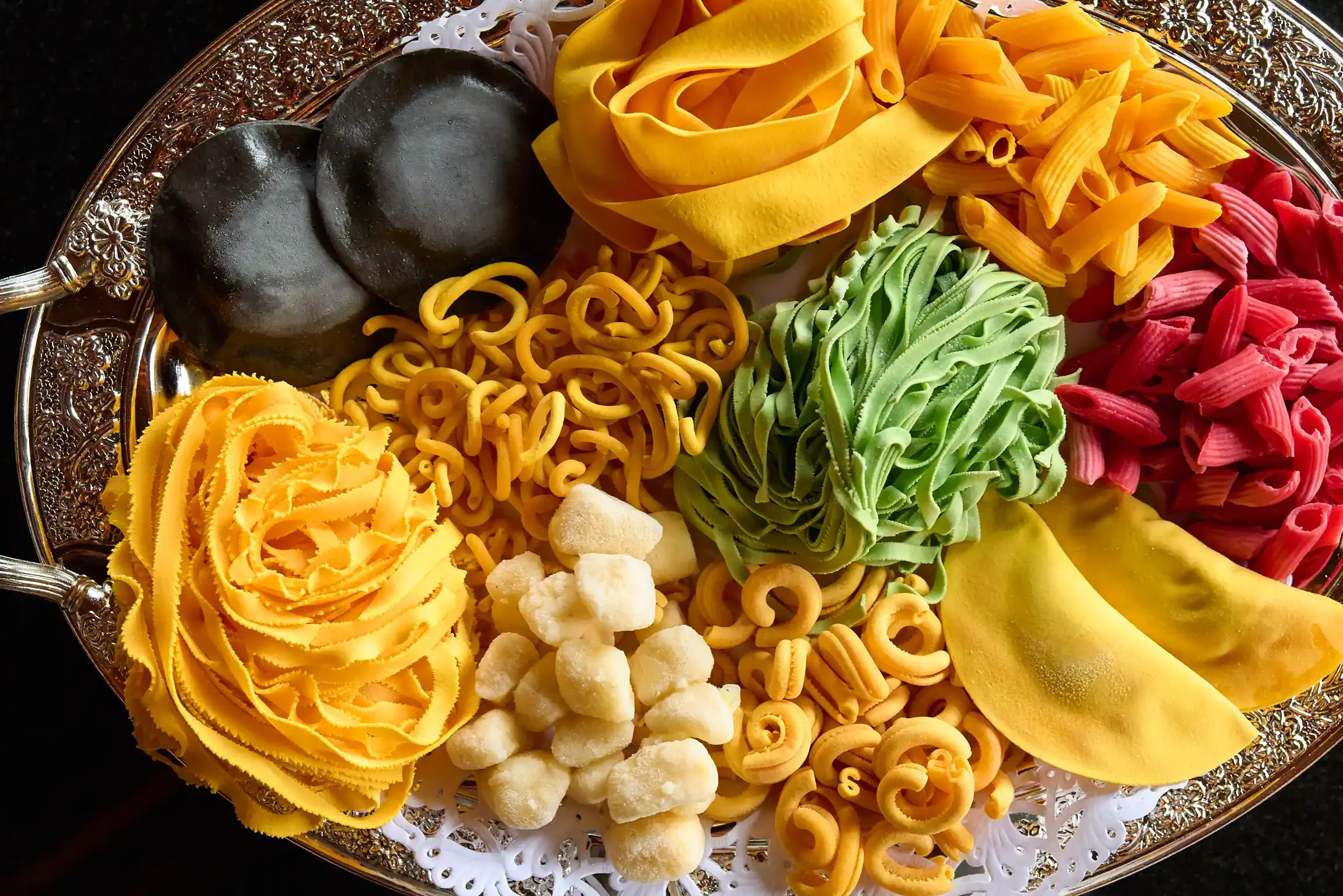 Display of various colorful uncooked pasta shapes on an ornate silver tray. By NJ editorial food photographer & food stylist team Bartholomew Studio.