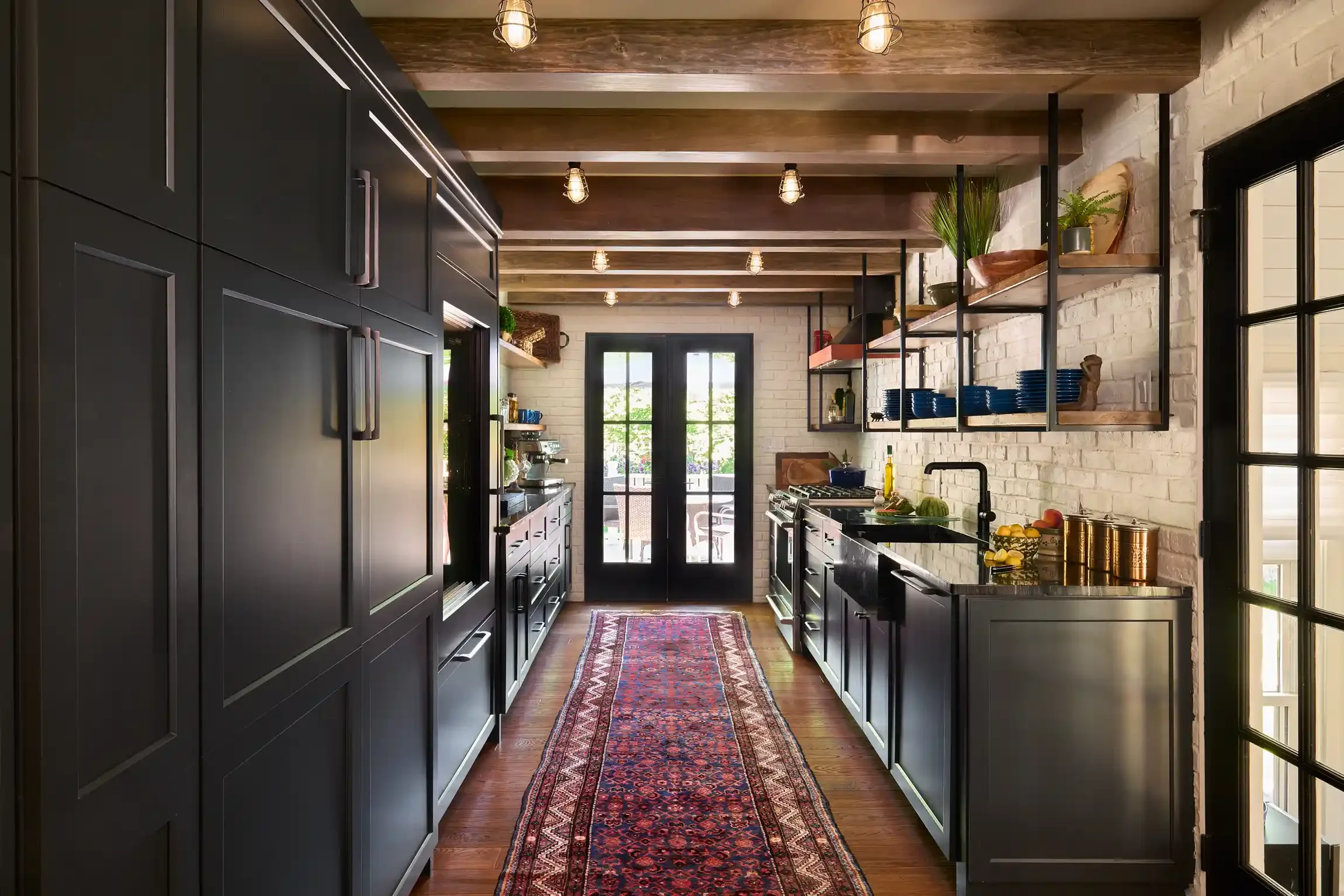 Modern kitchen with black cabinets, open wooden shelves, and a brick wall. French doors lead outside, and a colorful runner rug is on the wooden floor. By Philadelphia, Pennsylvania interior photographer Bartholomew Studio.