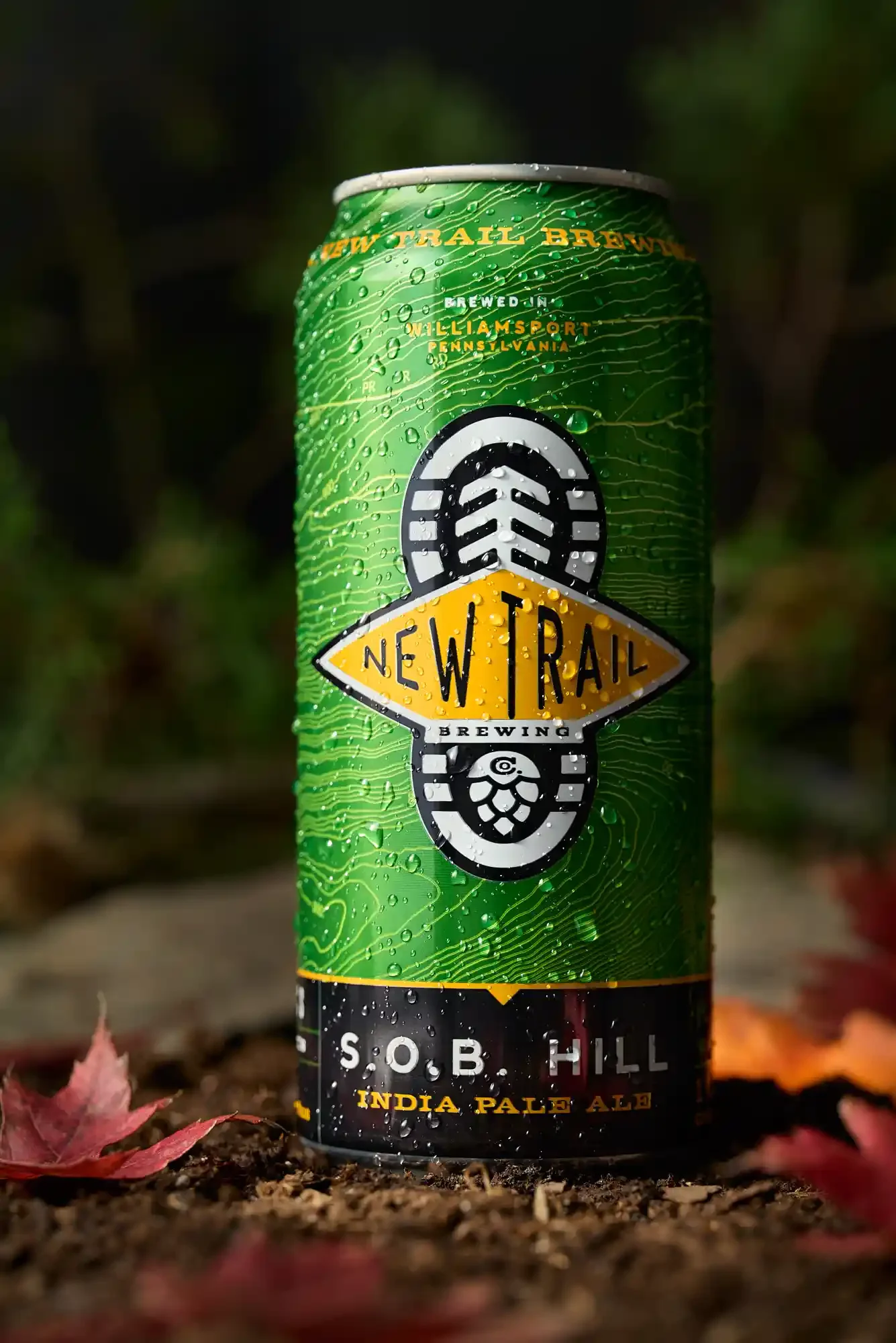 A green can of New Trail brewing S.O.B. Hill India Pale Ale with water droplets on the surface, placed on the ground with fallen leaves and a blurred outdoor background. By Philadelphia, PA beer photographer & drink stylist team Bartholomew Studio.