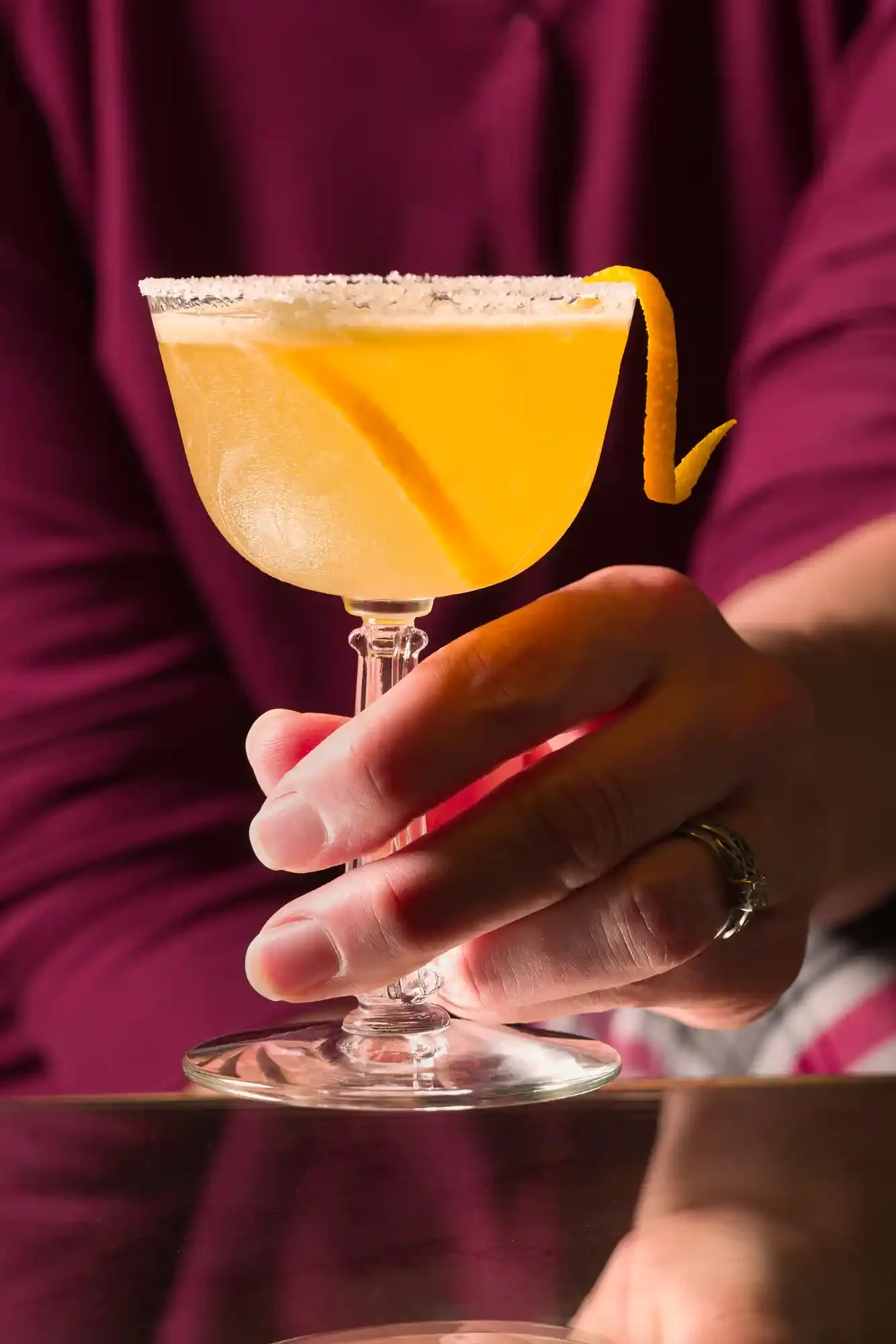 A hand holding a cocktail glass filled with a yellow drink, garnished with a lemon twist, against a dark background. By New Jersey drink photographer & drink stylist team Bartholomew Studio.