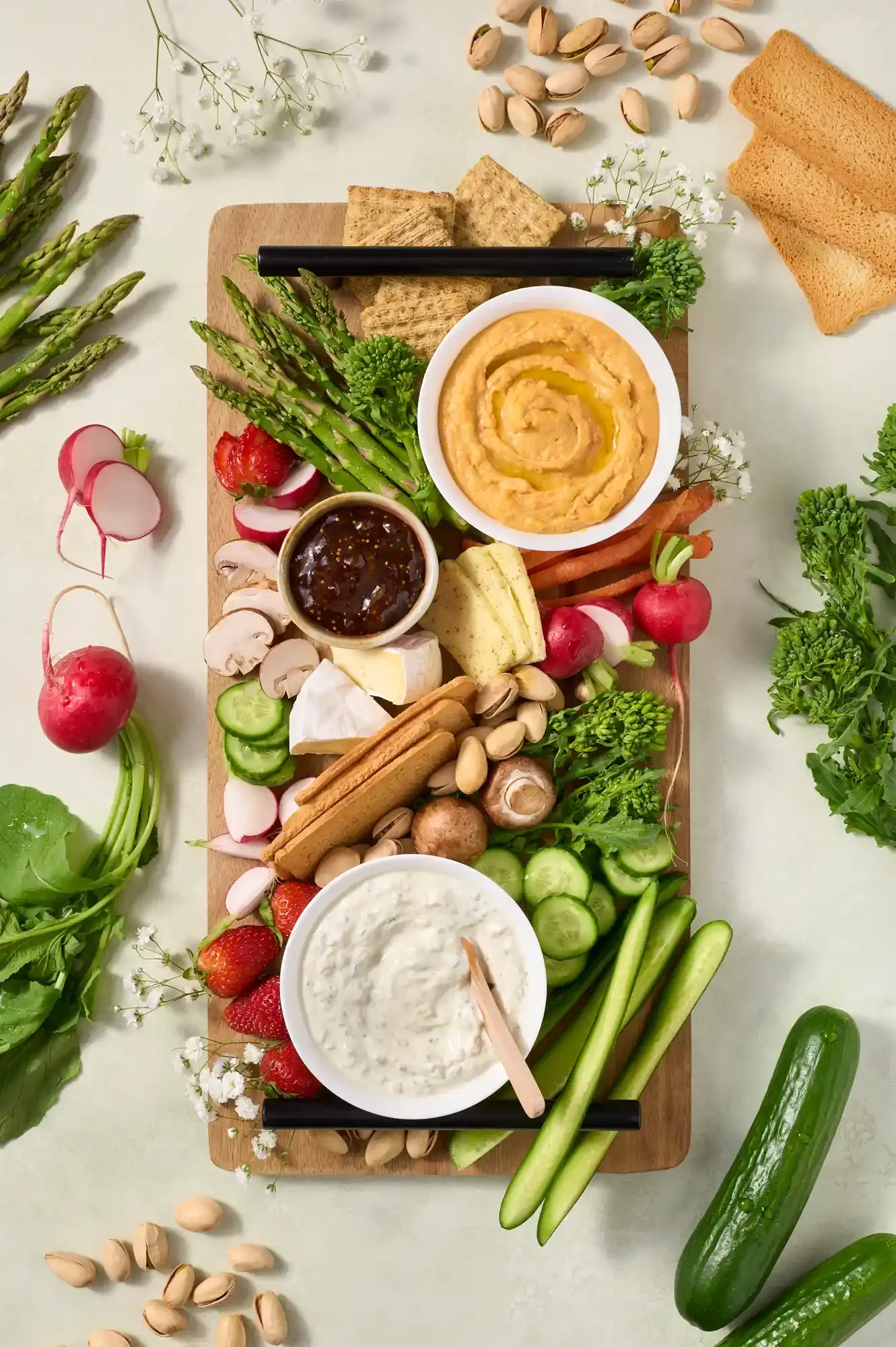 A wooden serving board holds various fresh vegetables, cheeses, crackers, and dips, including radishes, strawberries, cucumbers, mushrooms, pistachios, hummus, and dip, surrounded by fresh vegetables and herbs. By Philadelphia, PA food photographer.