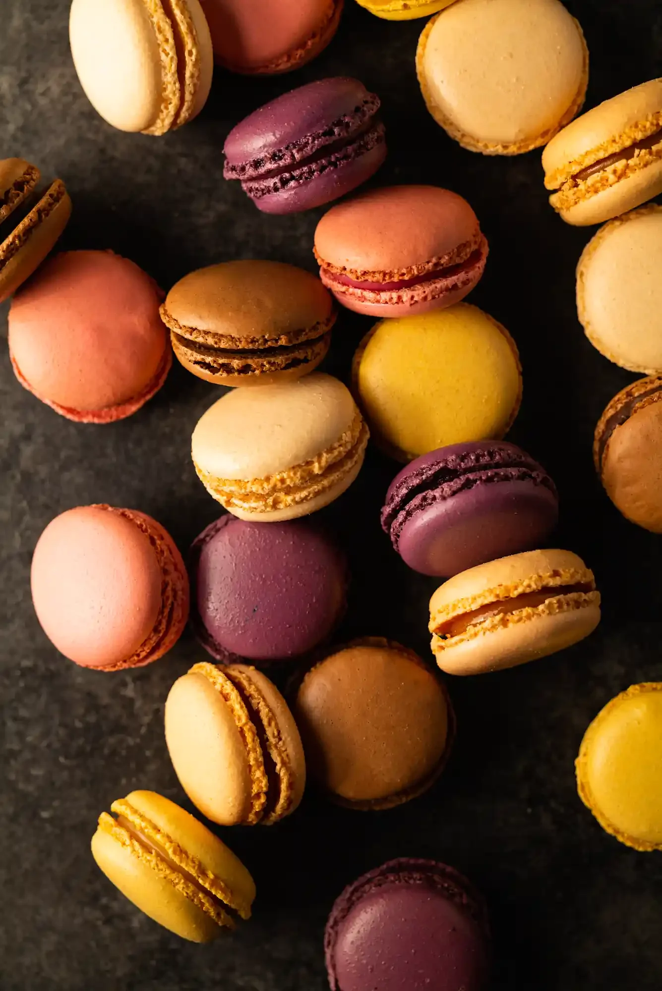 Colorful French macarons in pink, purple, yellow, white, and brown, arranged on a dark surface. By Philadelphia, PA food photographer & food stylist team Bartholomew Studio.