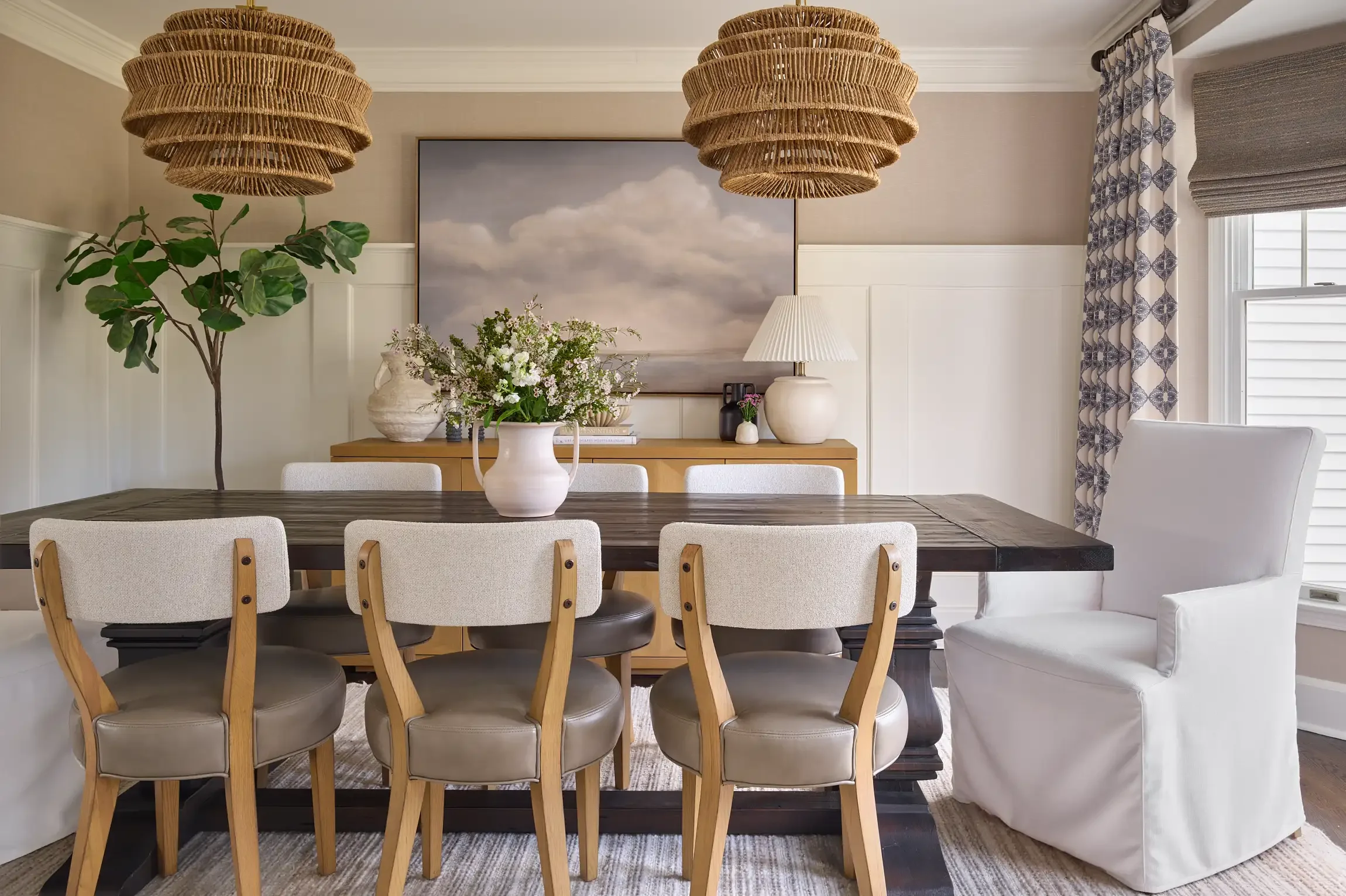 Dining room with a wooden table, beige chairs, a white vase with a floral arrangement, a sideboard with a lamp, artwork with clouds on the wall, a large green plant, and curtains with a blue pattern. By interior design photographer Paul Bartholomew.
