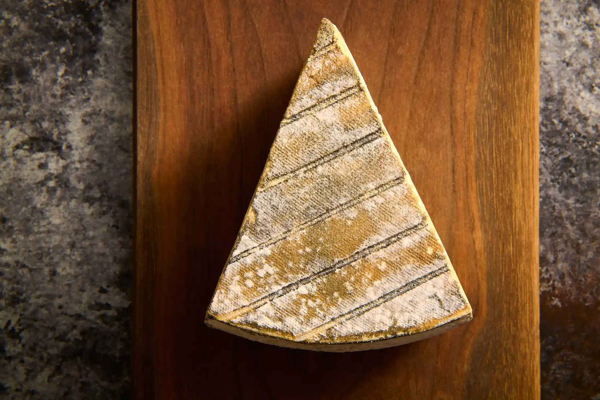A wedge of cheddar cheese with diagonal lines on top, on a wooden cutting board. By PA & NJ food photographer & food stylist team Bartholomew Studio.