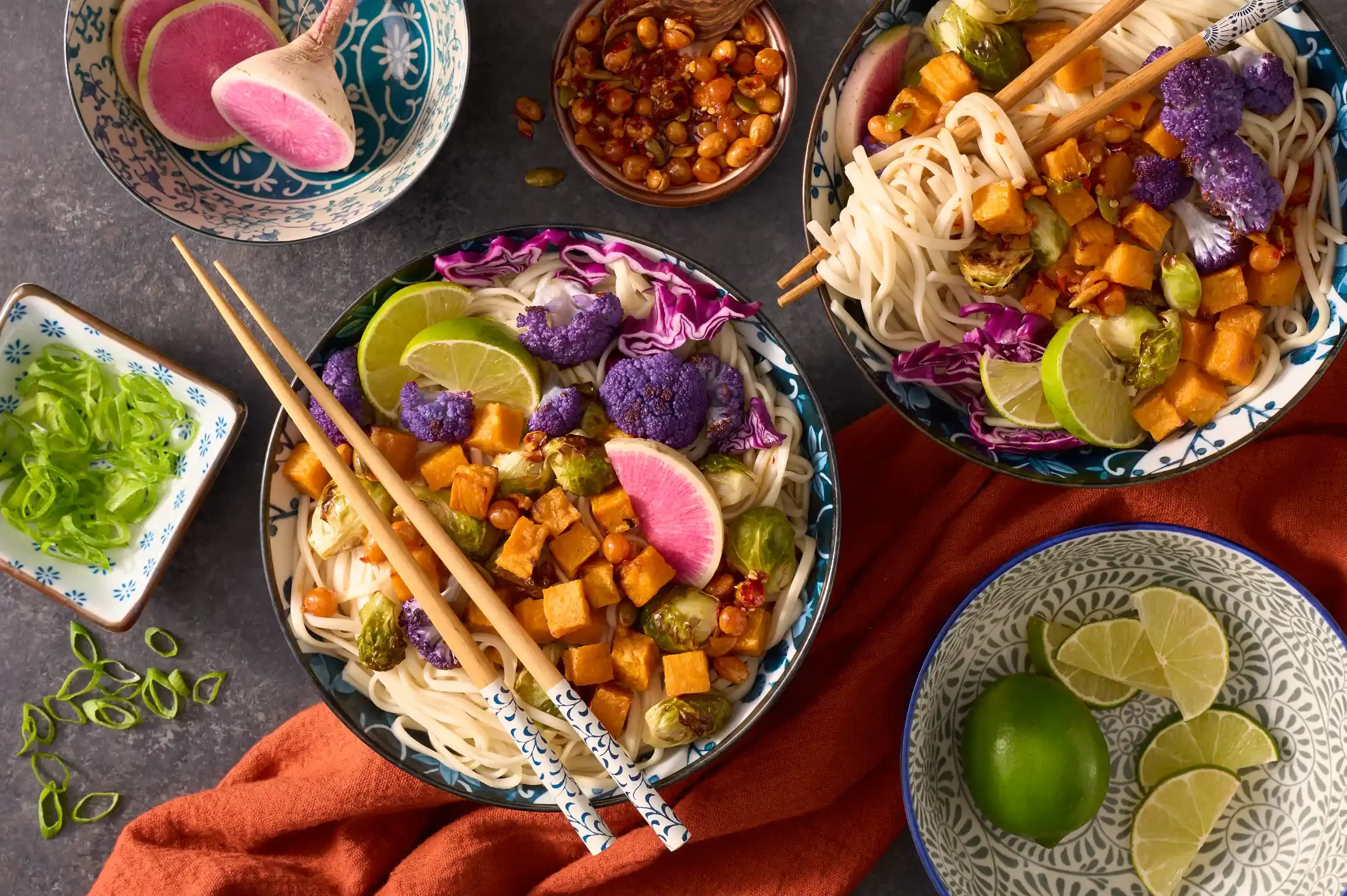Two bowls of Asian noodle salads with roasted vegetables, purple cauliflower, lime wedges, and radish slices. By Pennsylvania food photographer & food stylist team Bartholomew Studio.