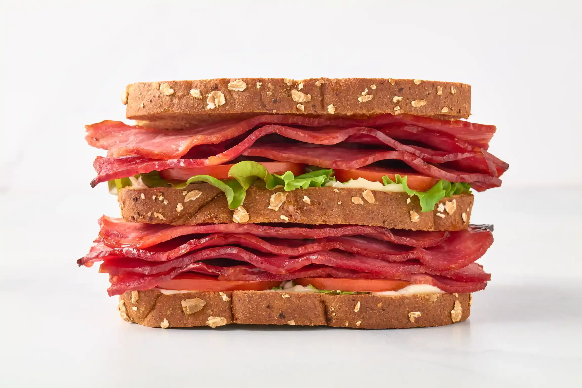 A close-up of a large BLT sandwich with multiple layers of bacon, lettuce, tomato, and whole grain bread slices. By Philadelphia, PA food photographer & food stylist team Bartholomew Studio.
