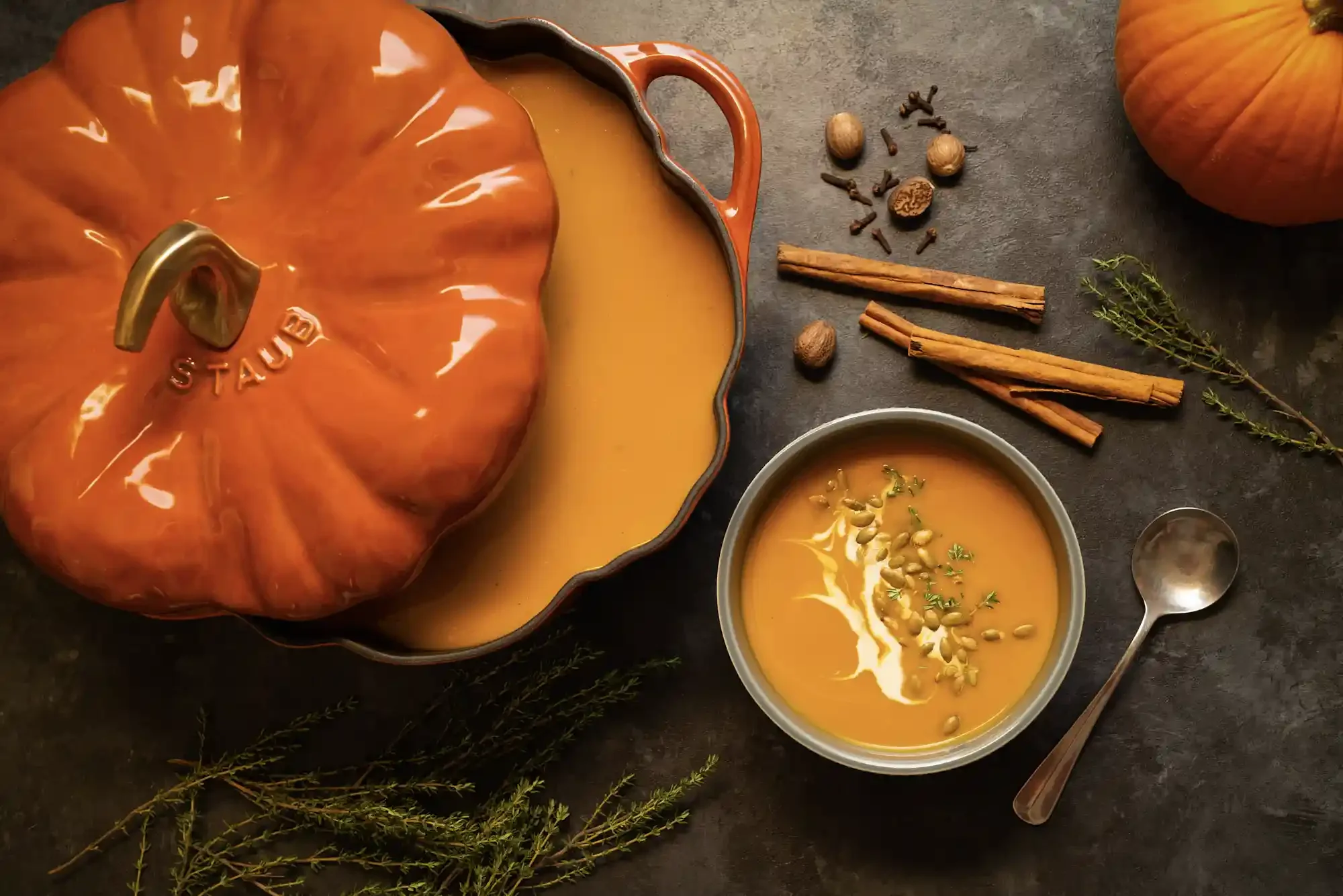 A pumpkin-shaped teapot filled with pumpkin soup, a bowl of pumpkin soup garnished with seeds, whole spices, and herbs, placed on a dark surface. By Philadelphia, PA food photographer & food stylist team Bartholomew Studio.