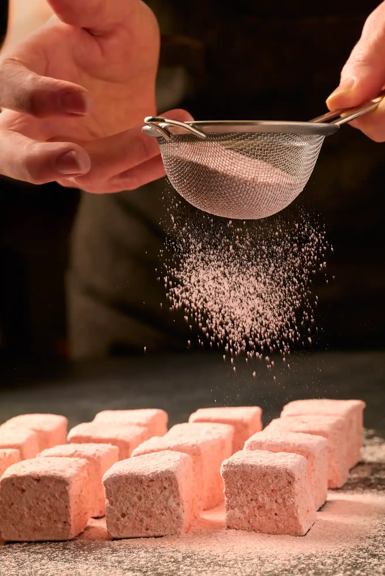 Pink powdered sugar being sifted through a metal sieve over pink marshmallows on a dark surface. By Pennsylvania food photographer & food stylist team Bartholomew Studio.