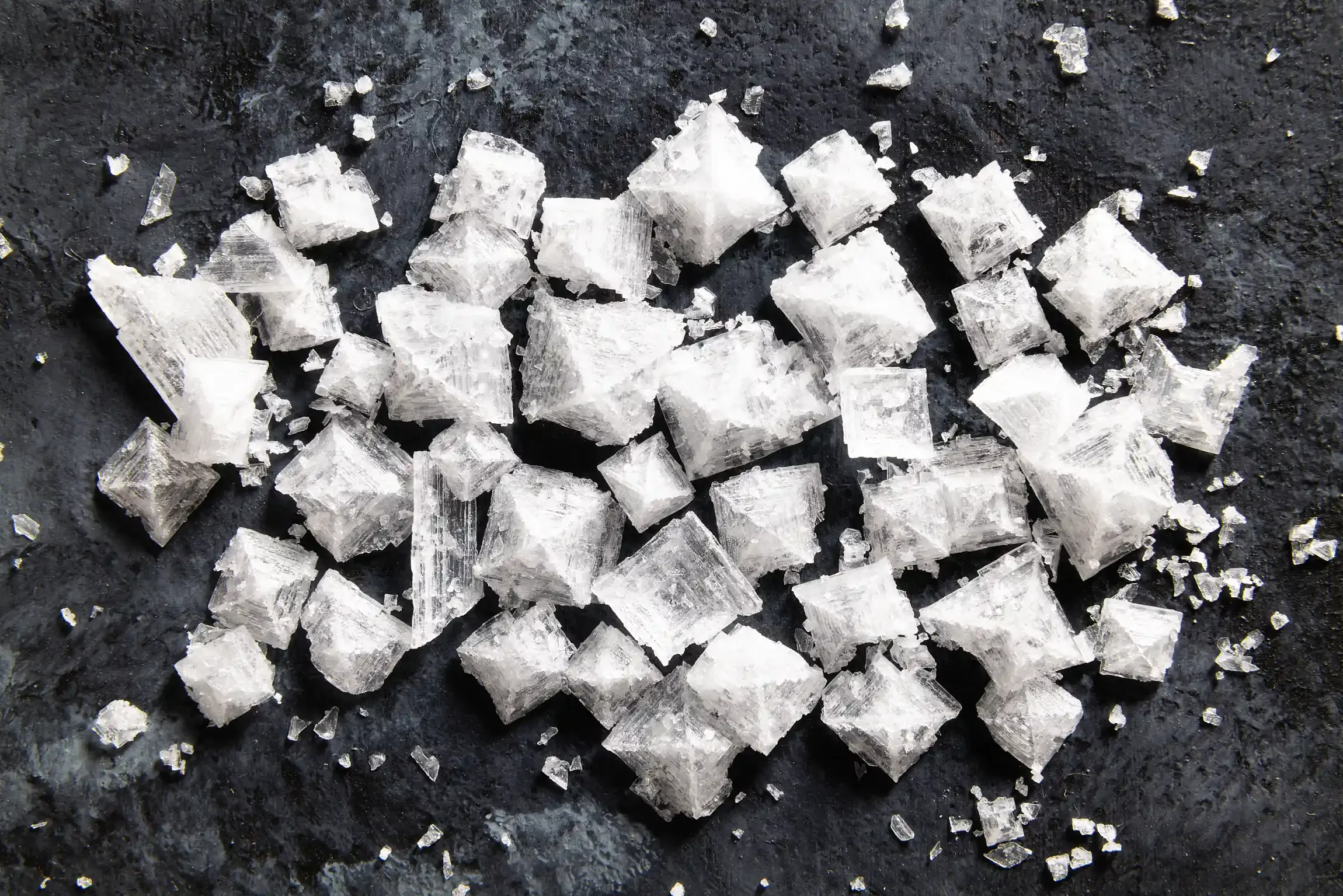 Chunks of coarse sea salt crystals scattered on a dark surface. By NYC, NY & PA food photographer & food stylist team Bartholomew Studio.