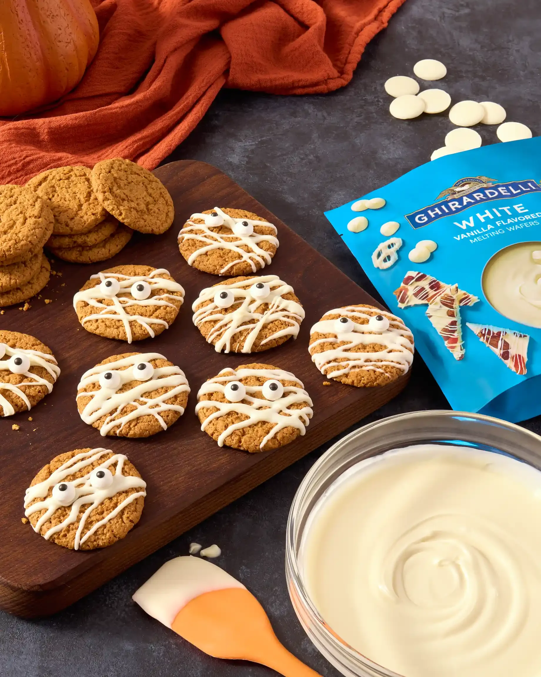 Halloween-themed mummy cookies decorated with white icing and candy eyes on a wooden board, a bag of white melting wafers, a bowl of white chocolate. By Philadelphia, PA & NJ food photographer & food stylist team Bartholomew Studio.