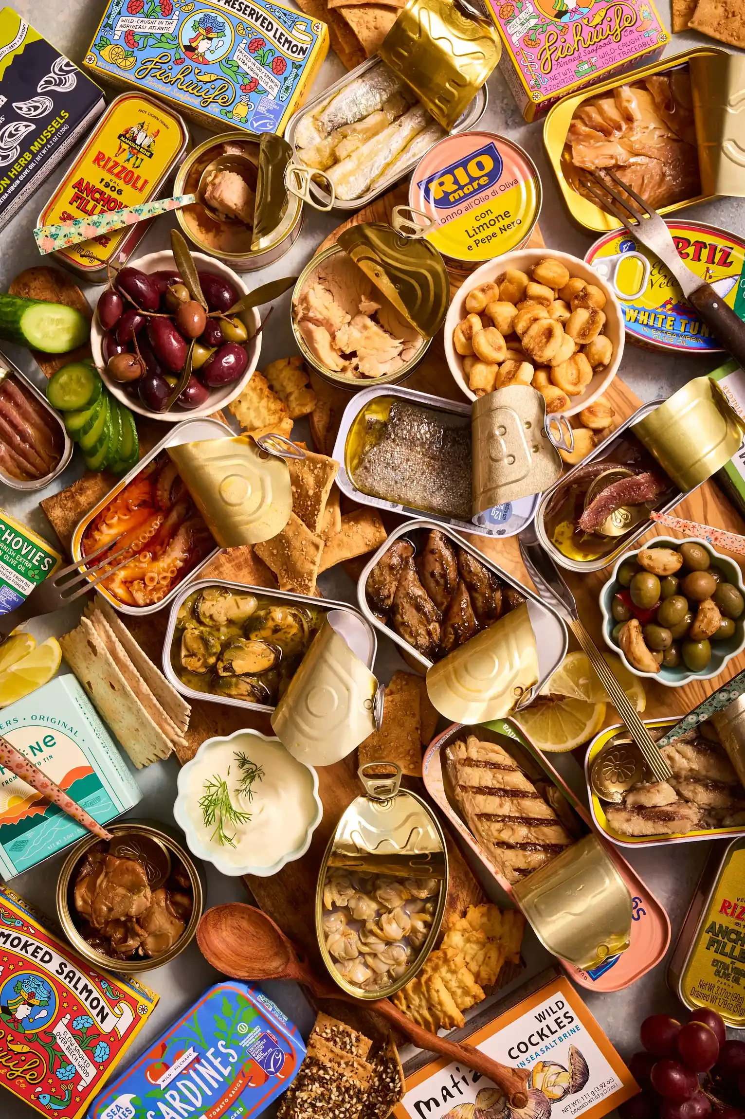 A variety of canned seafood and snacks on a table, including sardines, smoked salmon, oysters, anchovies, olives, and finger foods, with some fresh grapes and lemon slices. By PA, NJ & NYC food photographer & food stylist team Bartholomew Studio.