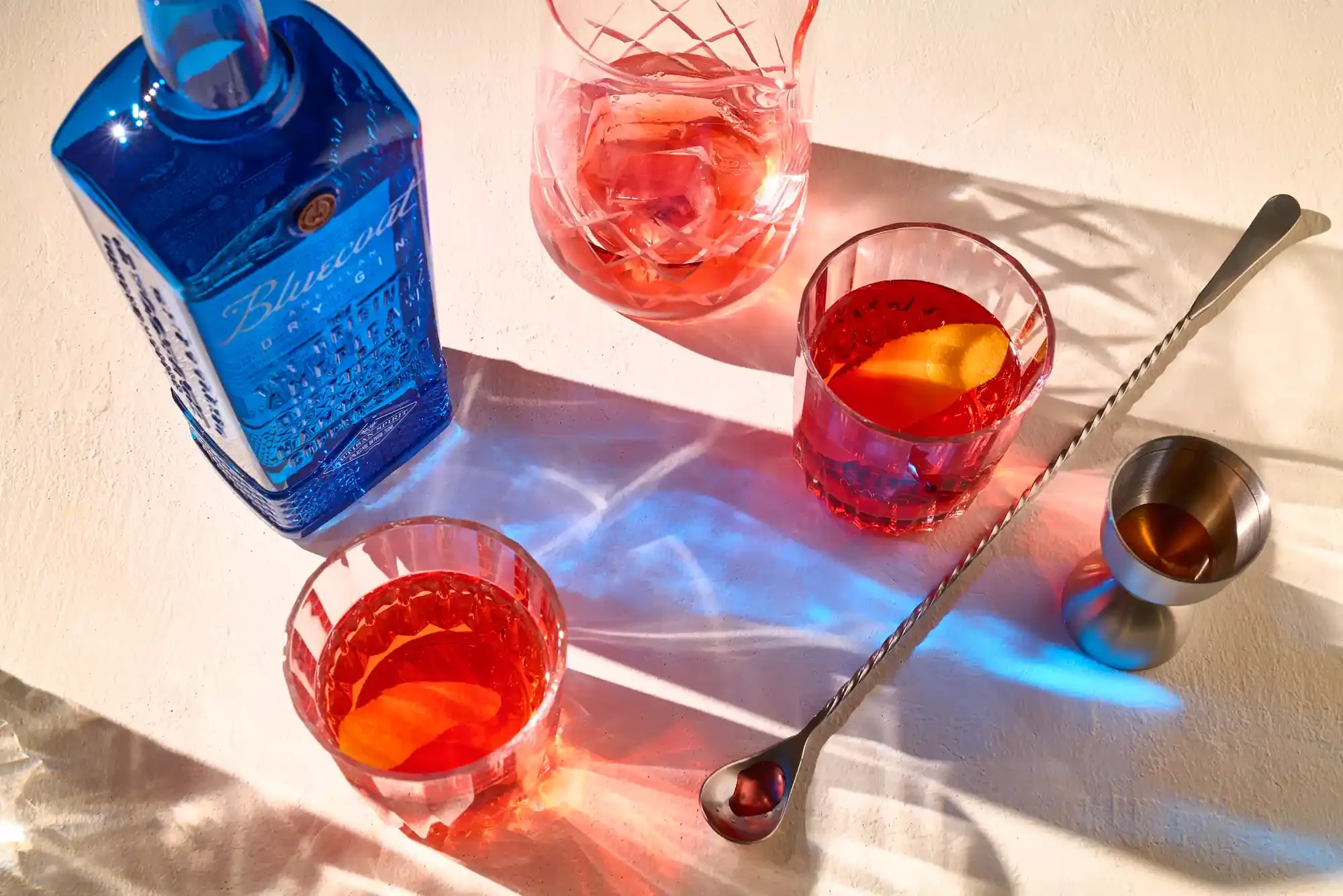 A bottle of Bluecoat gin, three glasses with Negroni drinks garnished with orange slices, and a cocktail shaker on a white surface with colorful shadows. By NJ & NYC drink photographer & drink stylist team Bartholomew Studio.