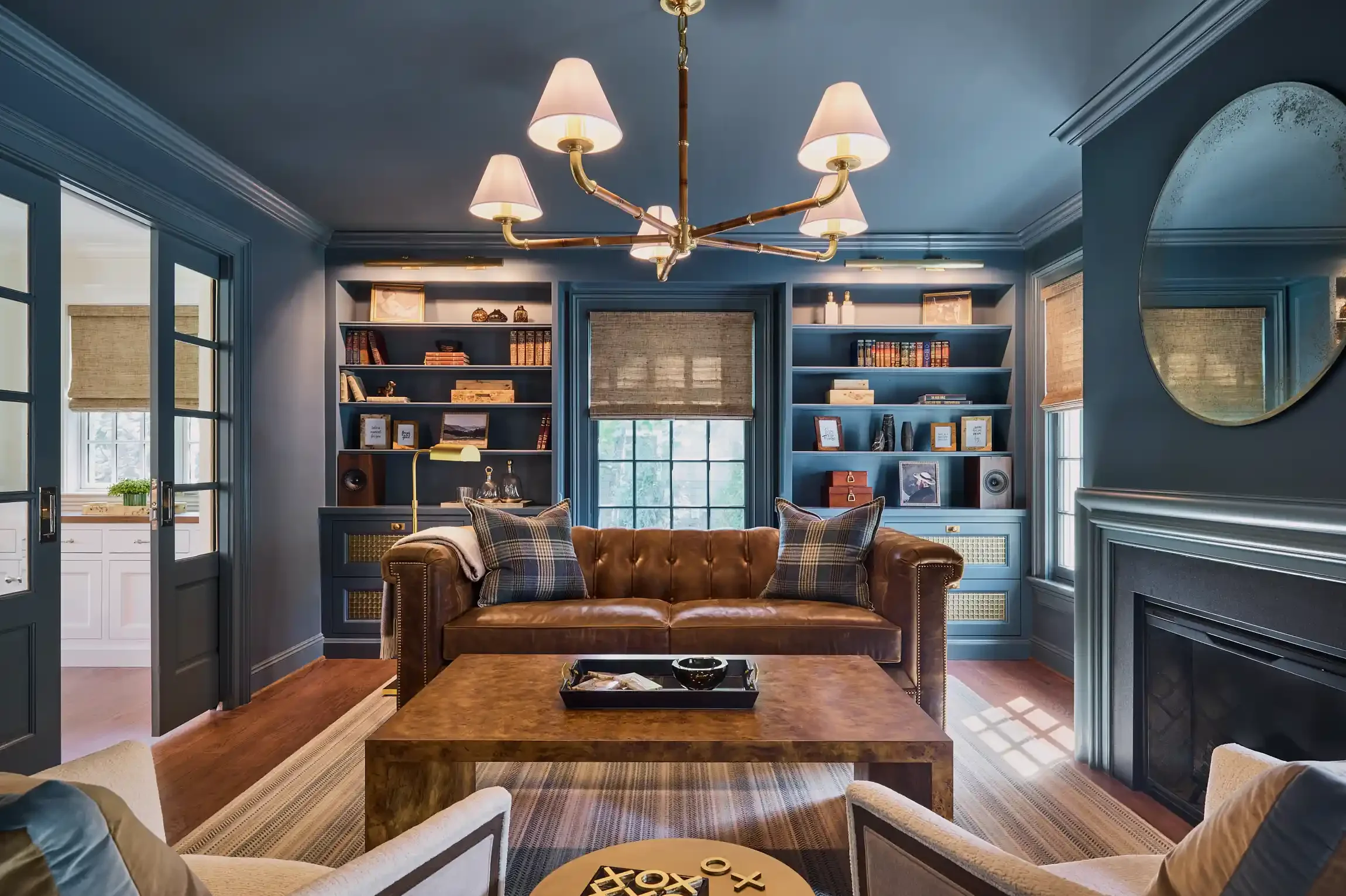 Living room with blue walls, a leather sofa with plaid pillows, a wooden coffee table, built-in blue bookshelves, a round mirror, and a modern chandelier. By NJ interior design photographer Bartholomew Studio.