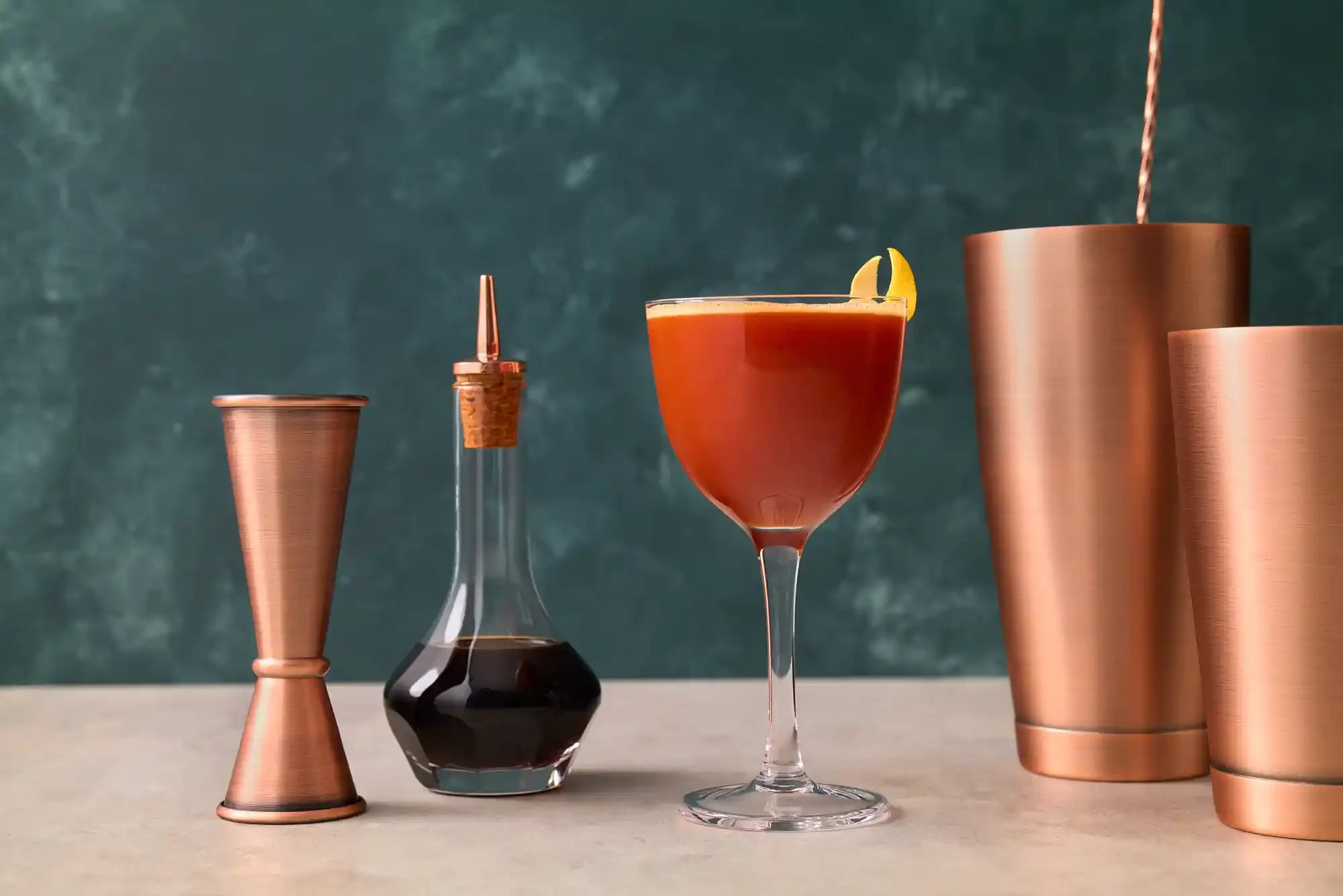 Trinidad Sour cocktail, garnished with a lemon wedge, beside copper-colored bar tools and containers on a light surface with a dark green textured background. By Philadelphia, PA cocktail photographer & cocktail stylist team Bartholomew Studio.