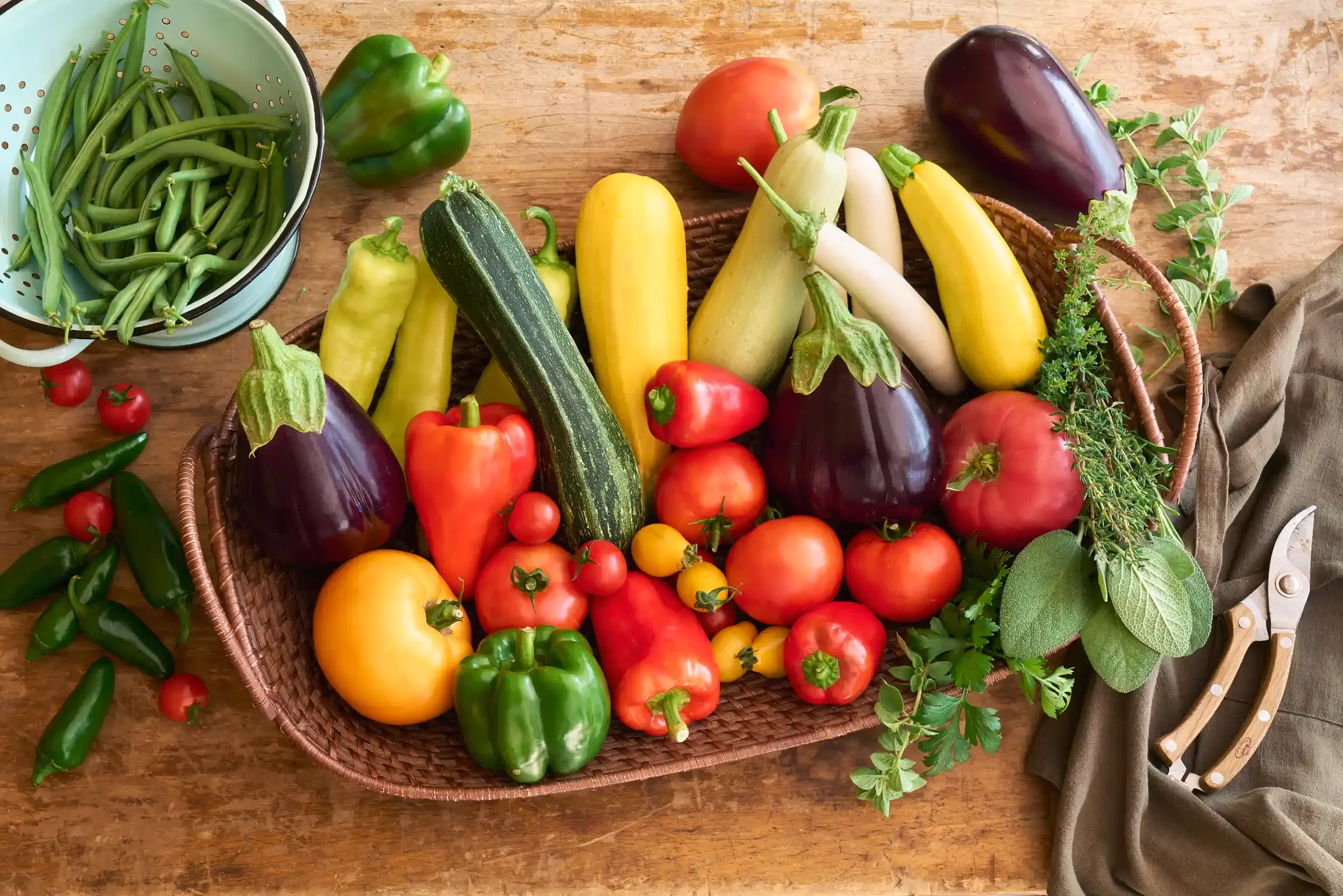 Basket of fresh vegetables including tomatoes, zucchini, eggplants, peppers, and herbs on a wooden table. By food photographer & food stylist team Bartholomew Studio.