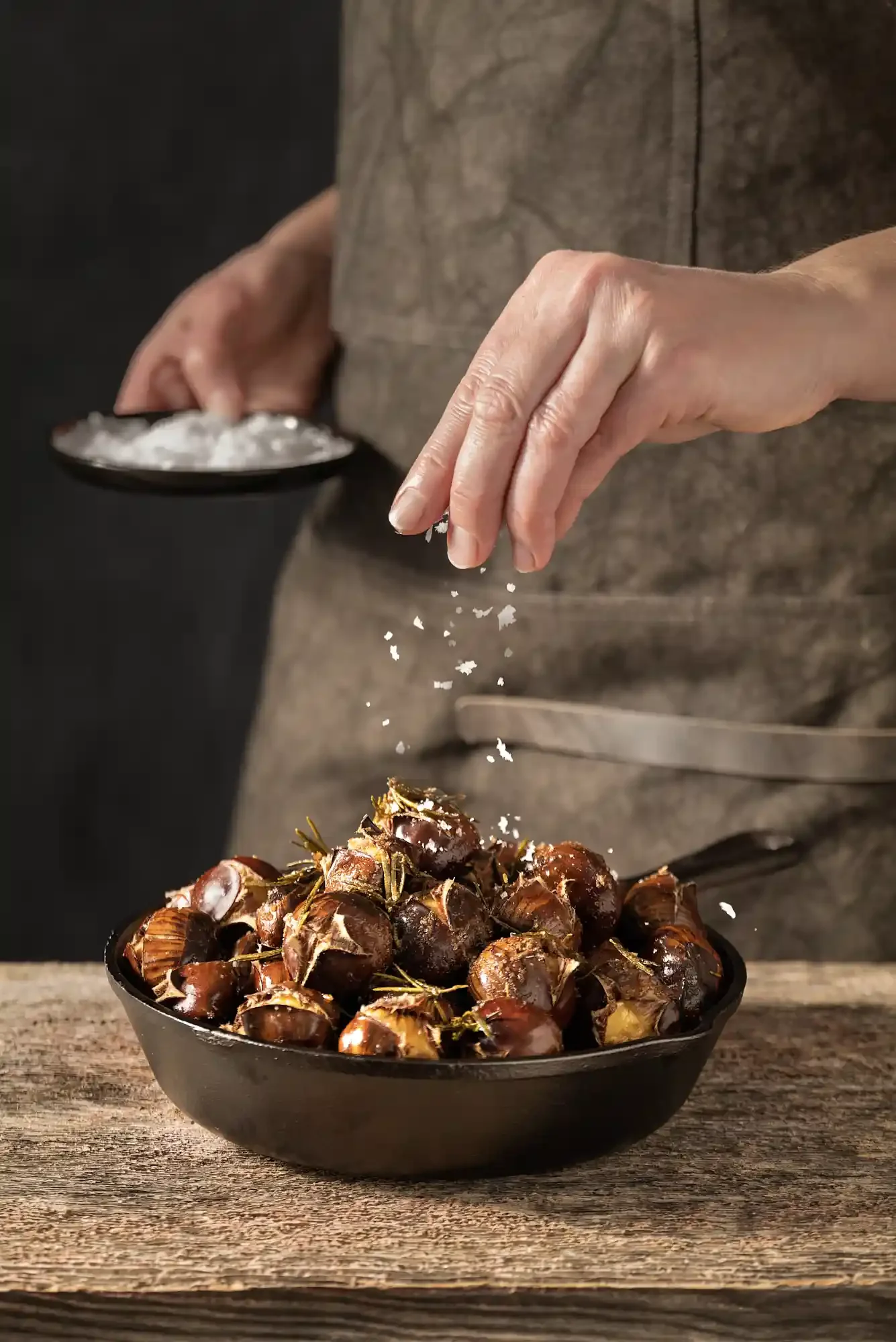 Person sprinkling salt on roasted chestnuts in a black bowl. By Philadelphia, PA food photographer & food stylist team Bartholomew Studio.