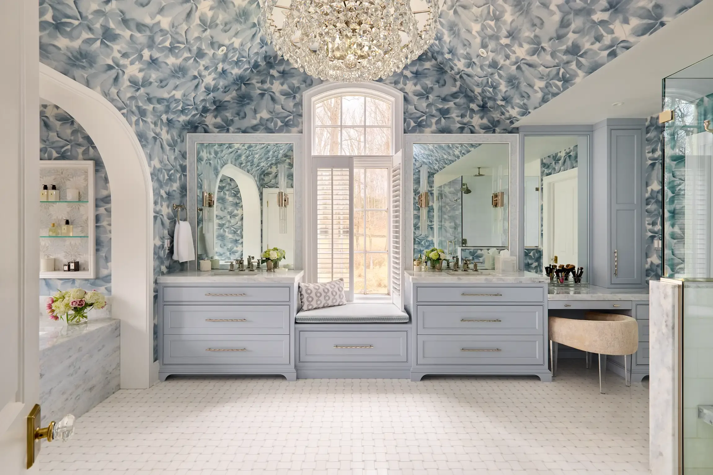 A luxurious bathroom with blue floral wallpaper on the ceiling and walls, featuring two large mirrors above white vanities with gold hardware. A crystal chandelier hangs from the ceiling, and a window with shutters allows natural light to brighten th
