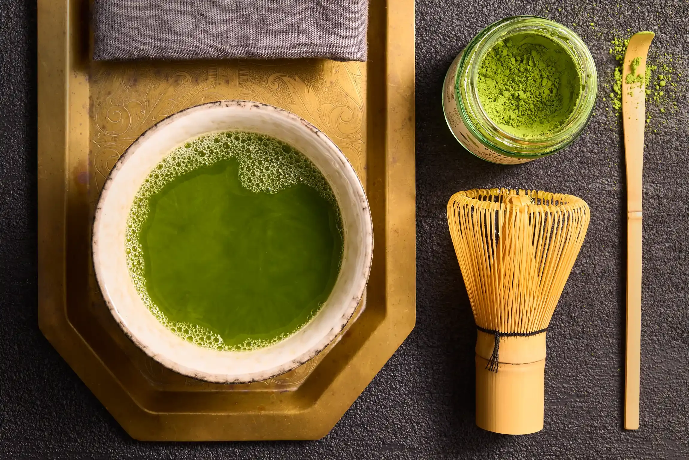 A traditional Japanese tea set with a bowl of matcha green tea, a container of matcha powder, a bamboo whisk, and a bamboo scoop on a dark surface. By Philadelphia, PA & New Jersey beverage photographer & beverage stylist team Bartholomew Studio.