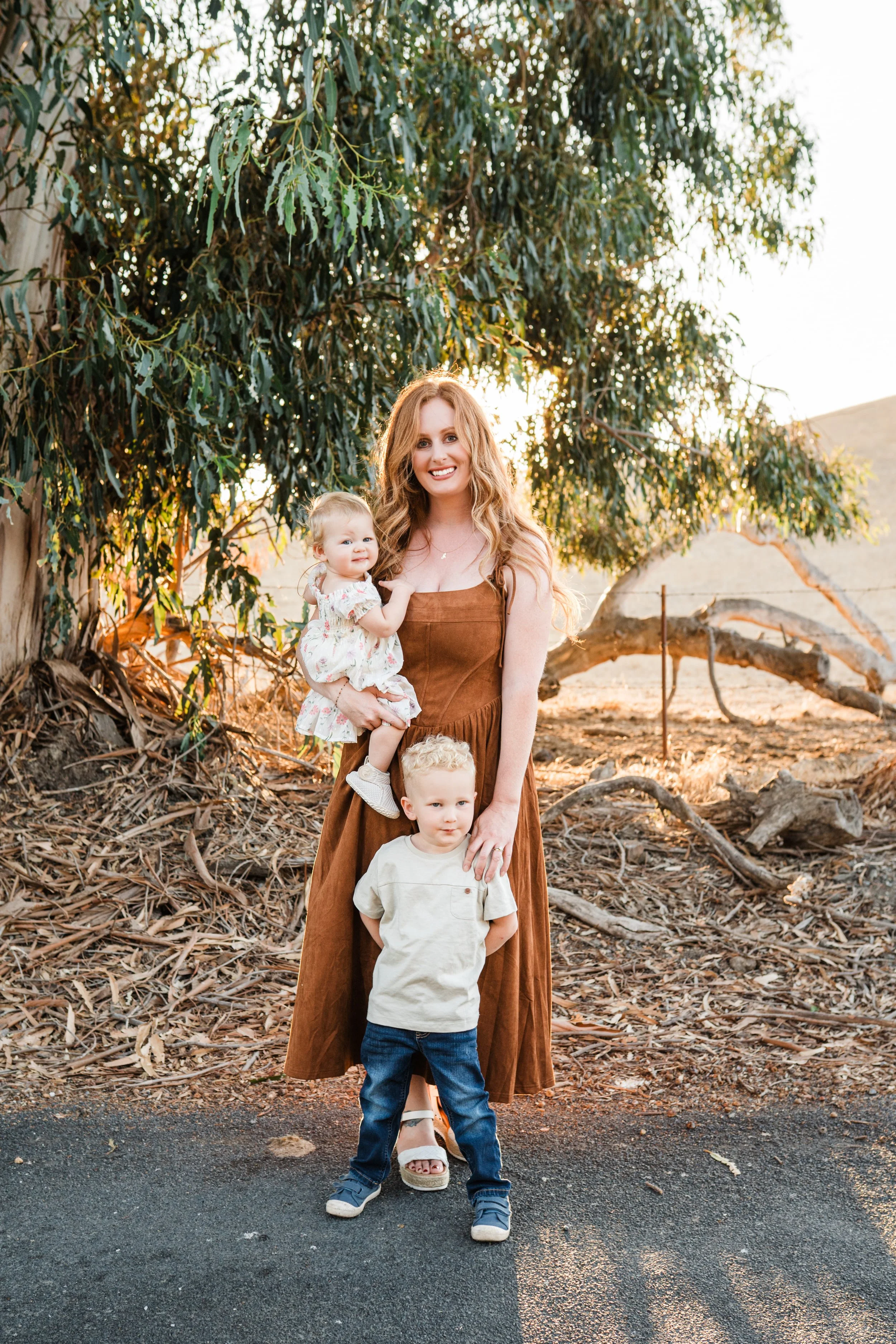 A woman with long, wavy red hair standing outdoors at sunset, holding a toddler girl and standing behind a young boy in front of a large tree with green leaves and a hillside.