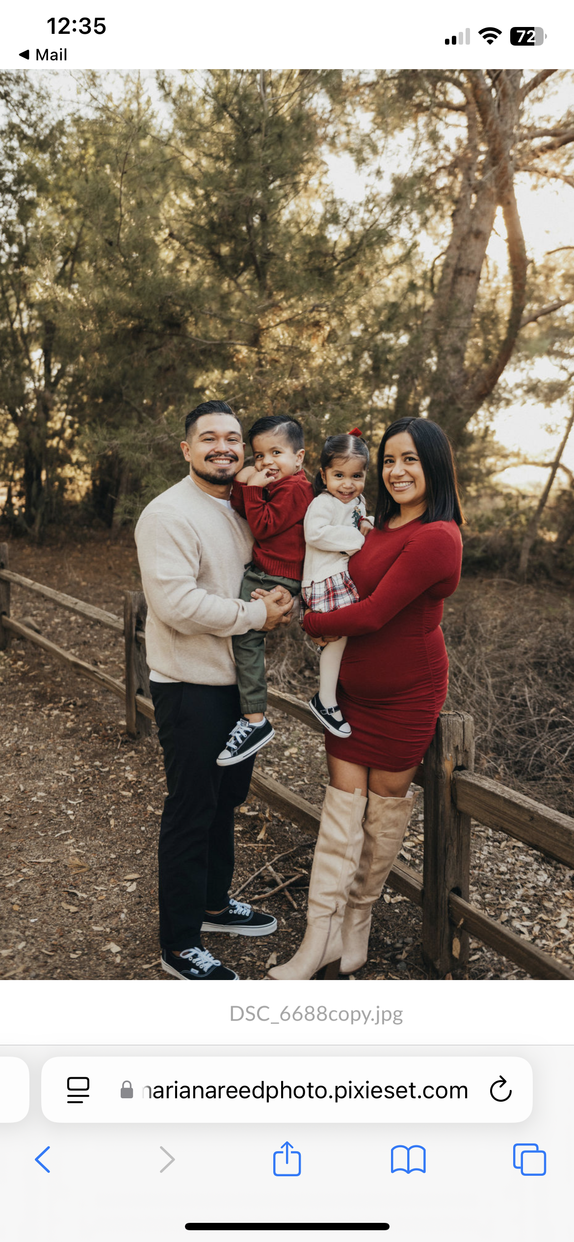 A family of four outdoors at sunset, standing near a wooden fence, smiling at the camera. The father, wearing a beige sweater, holds a young boy in a red sweater. The mother, in a red dress and cream over-the-knee boots, holds a young girl in a white sweater and plaid skirt. Trees are in the background.