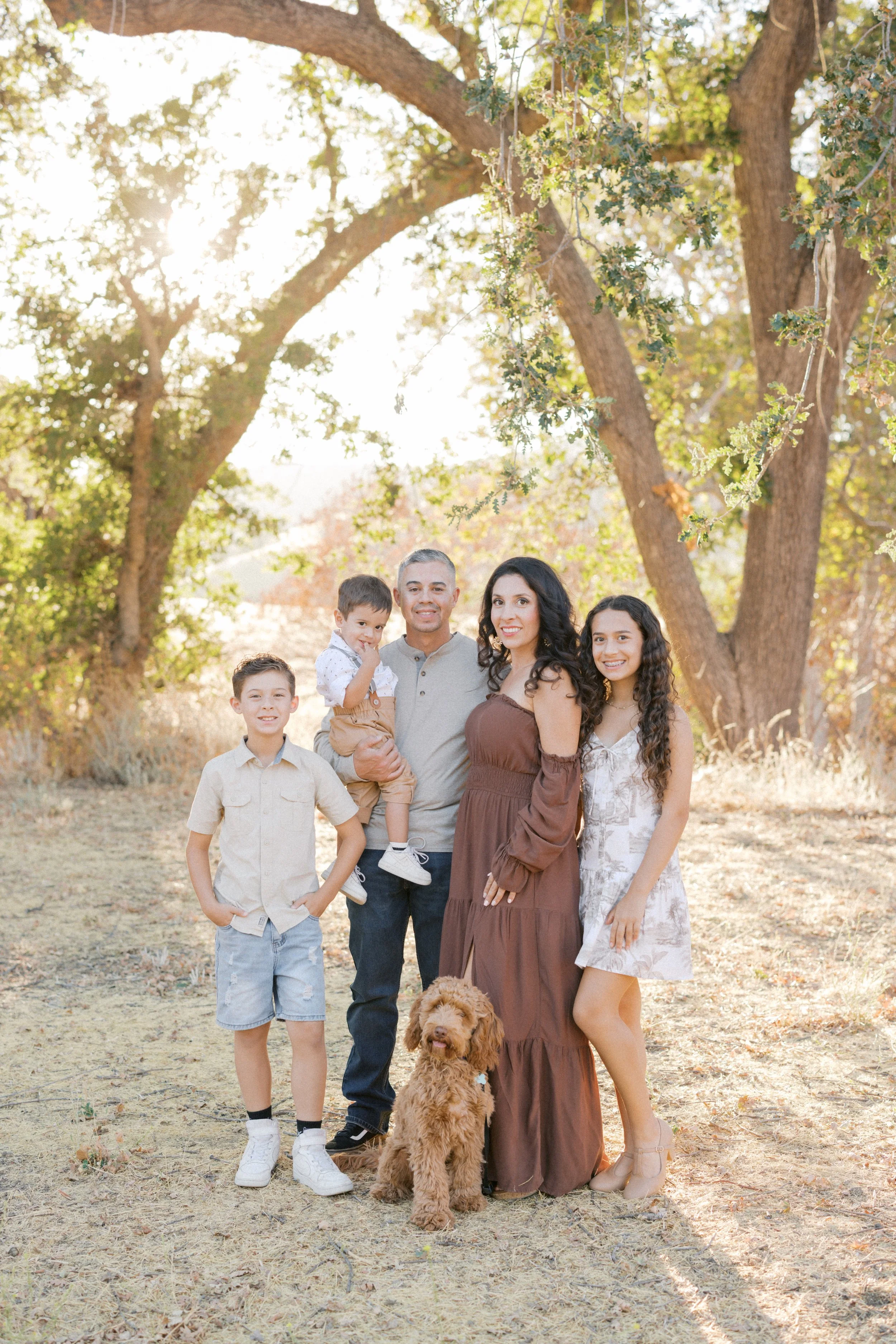 Family of five with dog standing outdoors in a park during fall, with tall trees and sunlight filtering through leaves in the background.