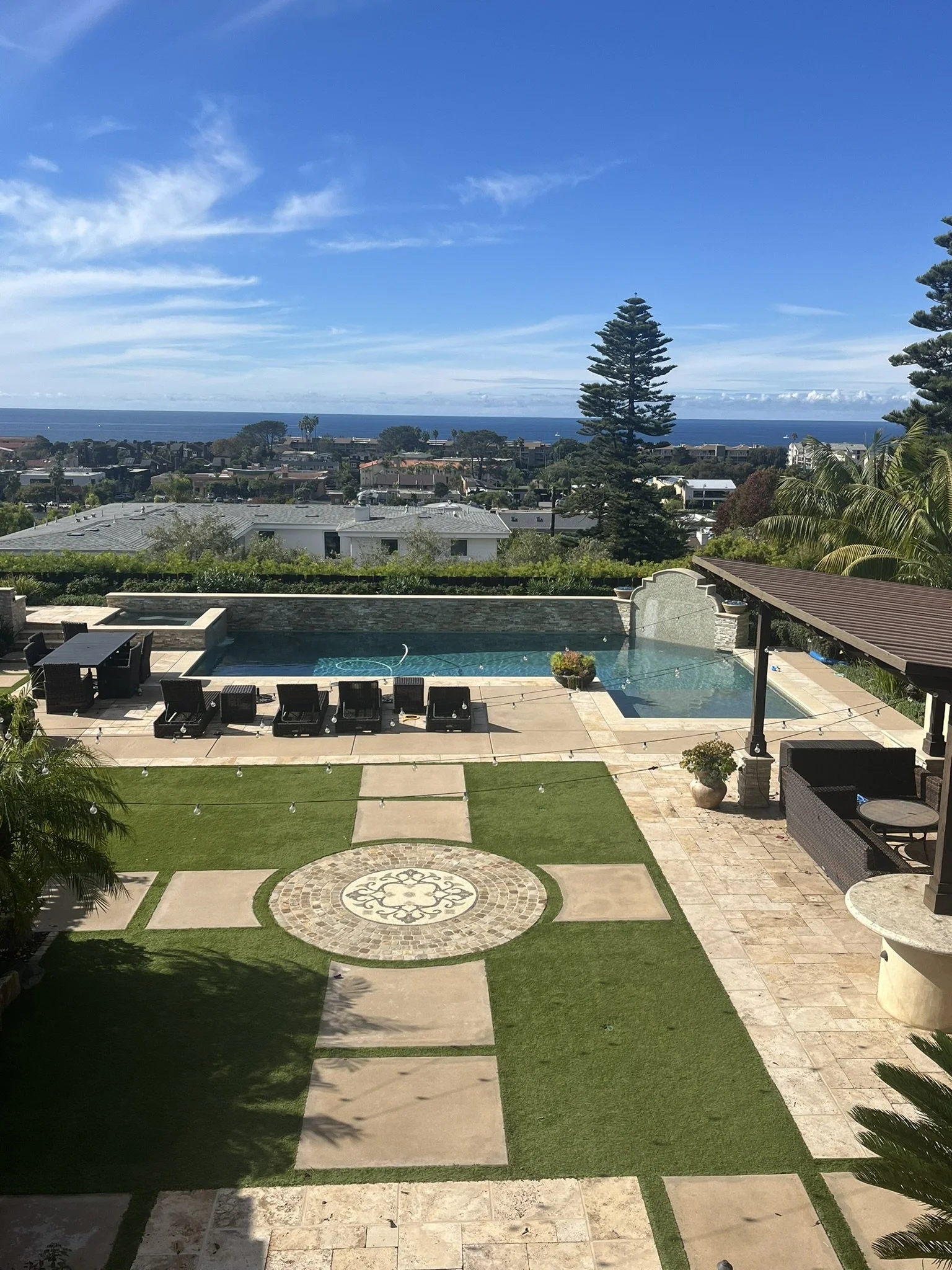 A backyard with a swimming pool, concrete patio, and outdoor furniture, overlooking a residential neighborhood and the ocean beyond under a blue sky.