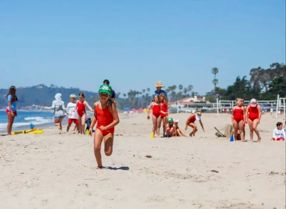 Kids playing volleyball and running on a sandy beach with ocean and palm trees in the background on a sunny day.