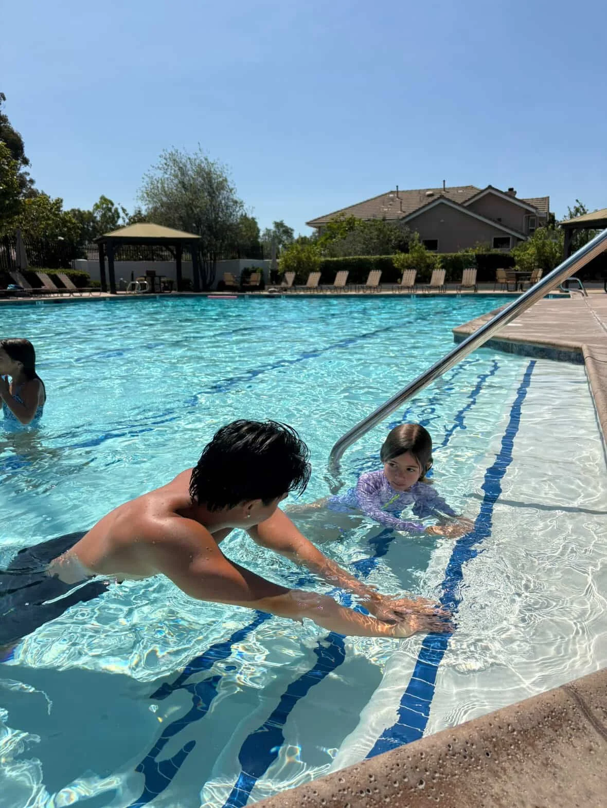 A backyard swimming pool scene with a man and a young girl near the pool steps, other children swimming, lounge chairs, a covered pavilion, and houses in the background under a clear blue sky.
