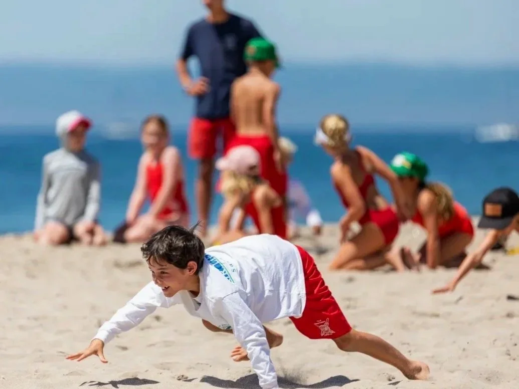 A young boy in a white shirt and red shorts is crawling on the sand at the beach, with a group of kids and an adult in the background, some kneeling and some standing, near the water.