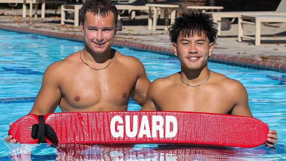 Two young male lifeguards standing in a swimming pool, holding a red rescue float with the word "GUARD" on it, smiling at the camera.