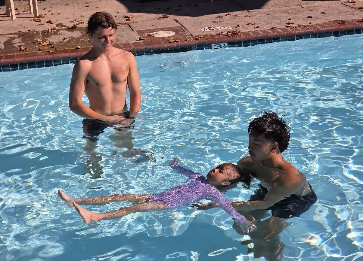 A young girl in a purple swimsuit floating on her back in a swimming pool, with two young men standing nearby, one holding her and the other standing in the water. The scene is outdoors on a sunny day.